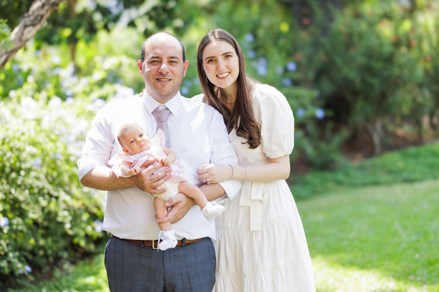 A couple poses with an infant in a sunny garden setting.