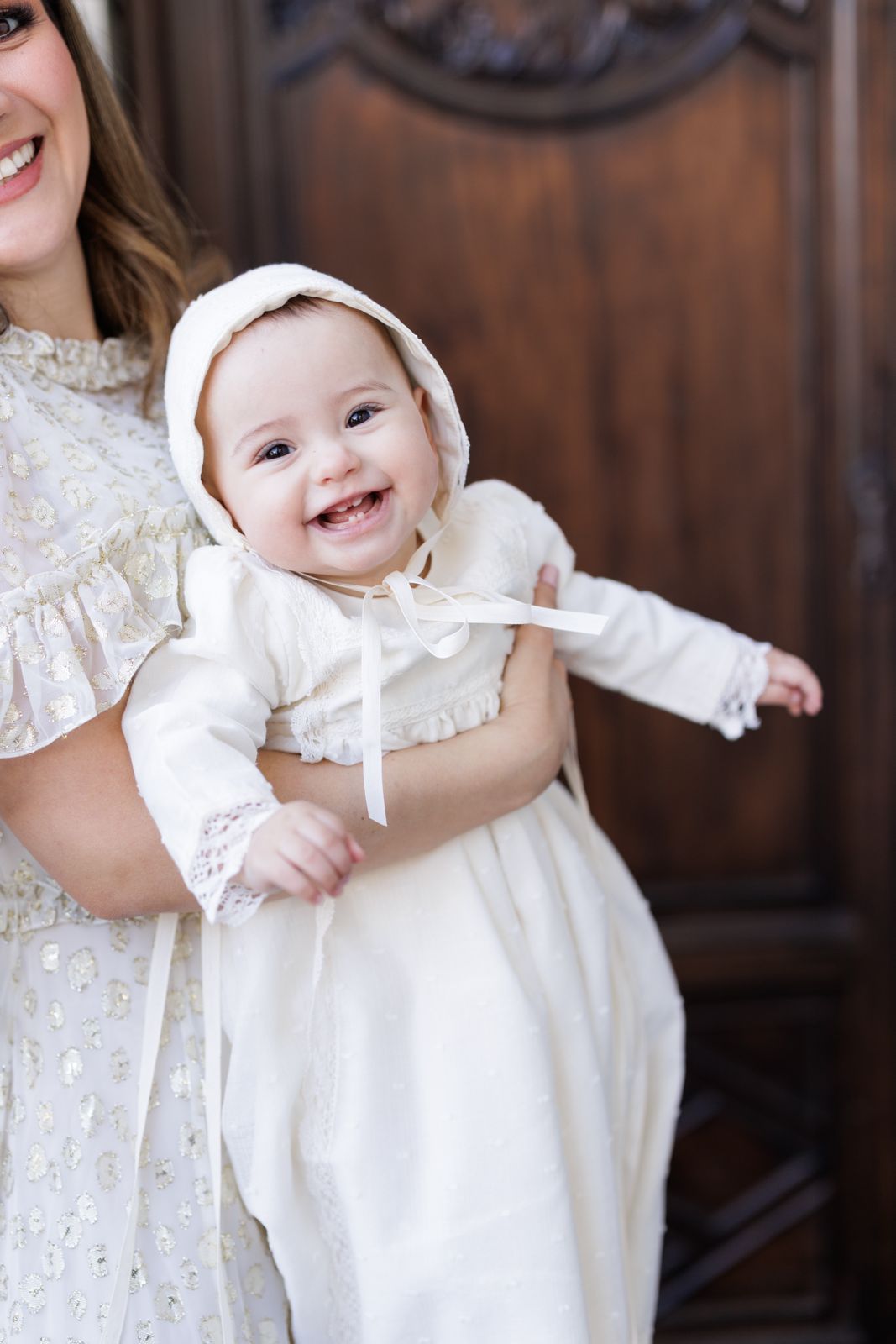 Baby wearing white christening gown and bonnet smiles while being held.