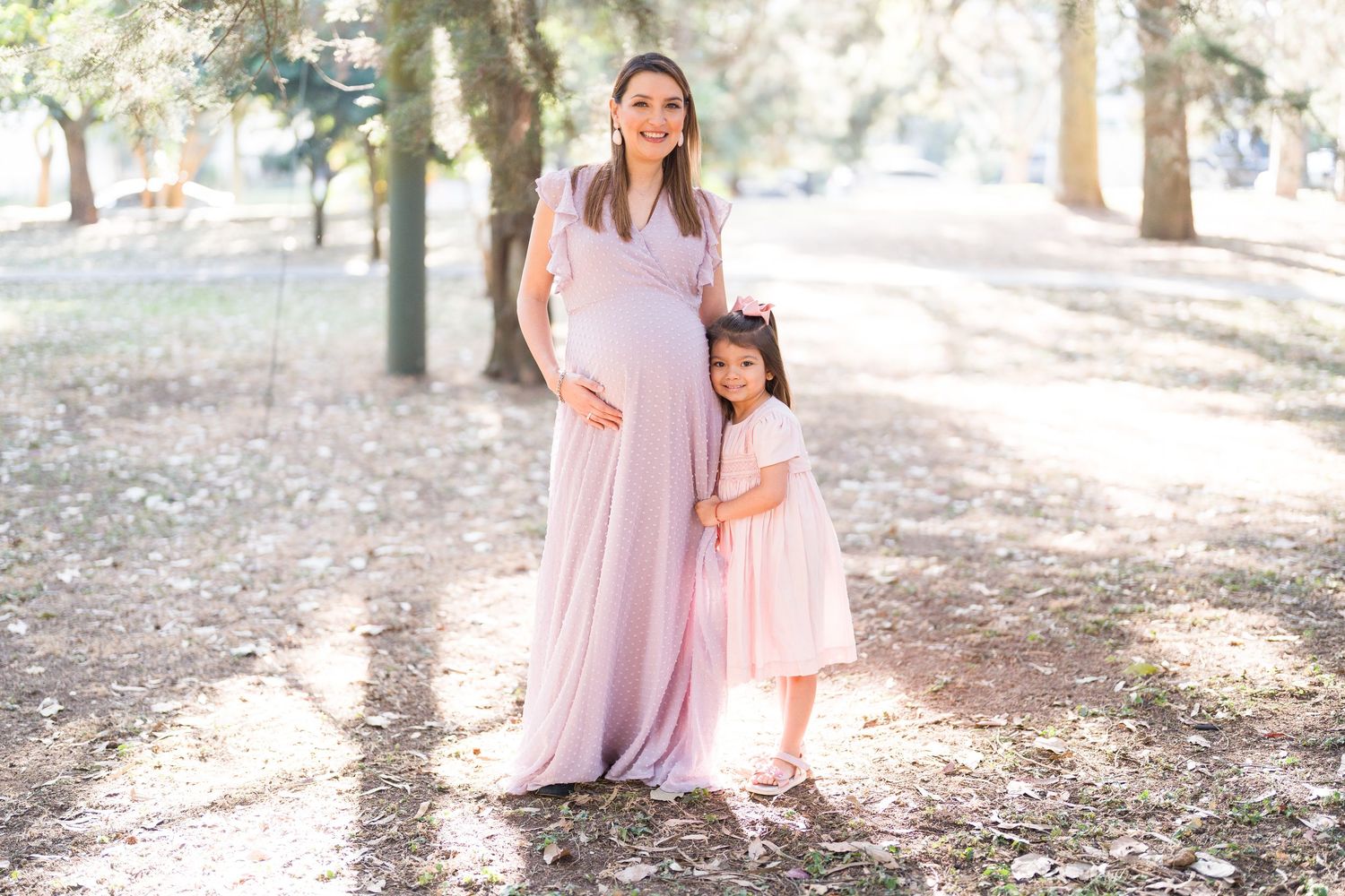 Mother and daughter in matching pink dresses share a moment in a forest setting.