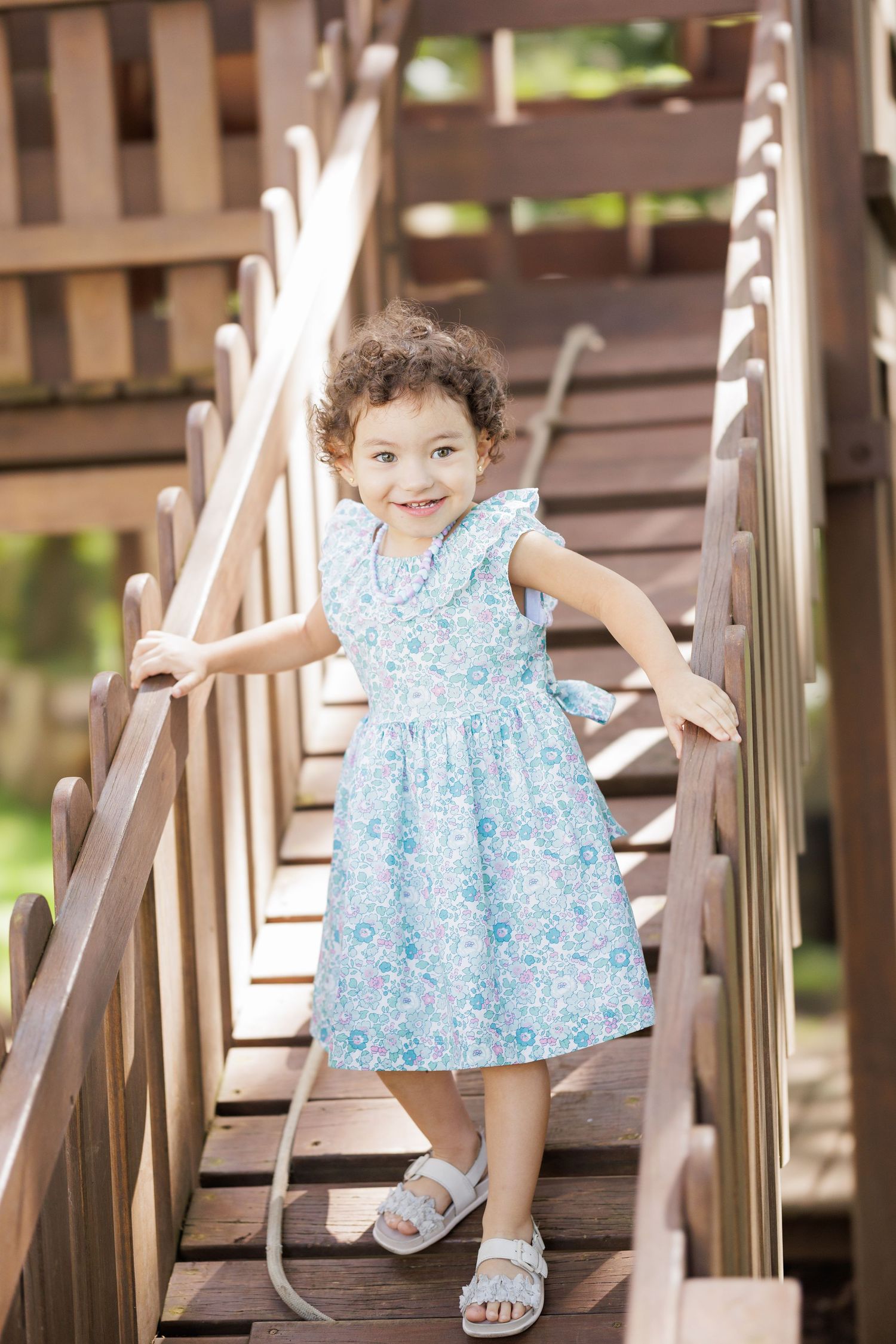 A child wearing a light blue floral dress stands on wooden stairs outdoors.