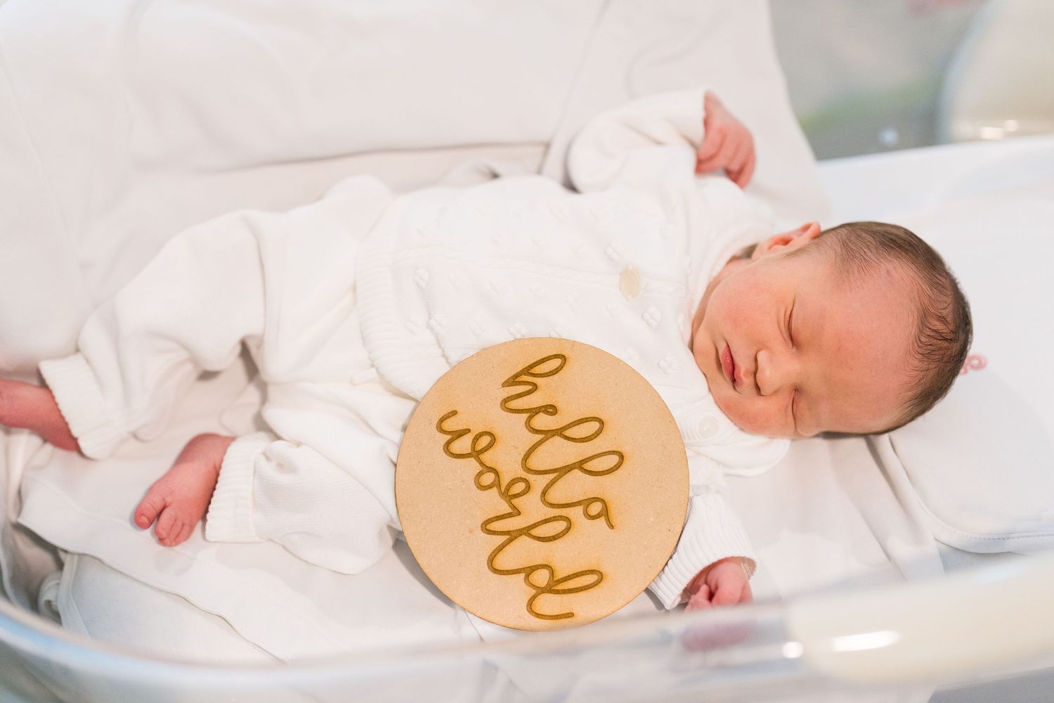 Newborn baby sleeping next to wooden hello sign on white bedding.