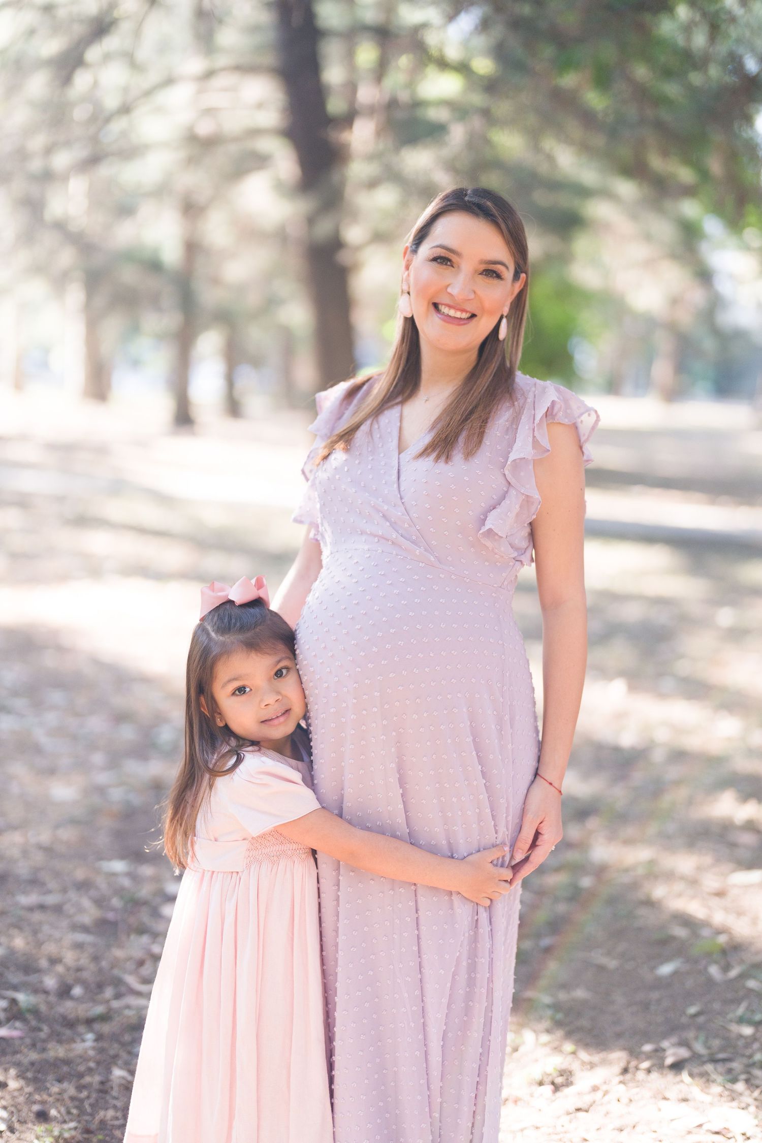 Mother and daughter embrace during outdoor maternity photo session in pink dresses.
