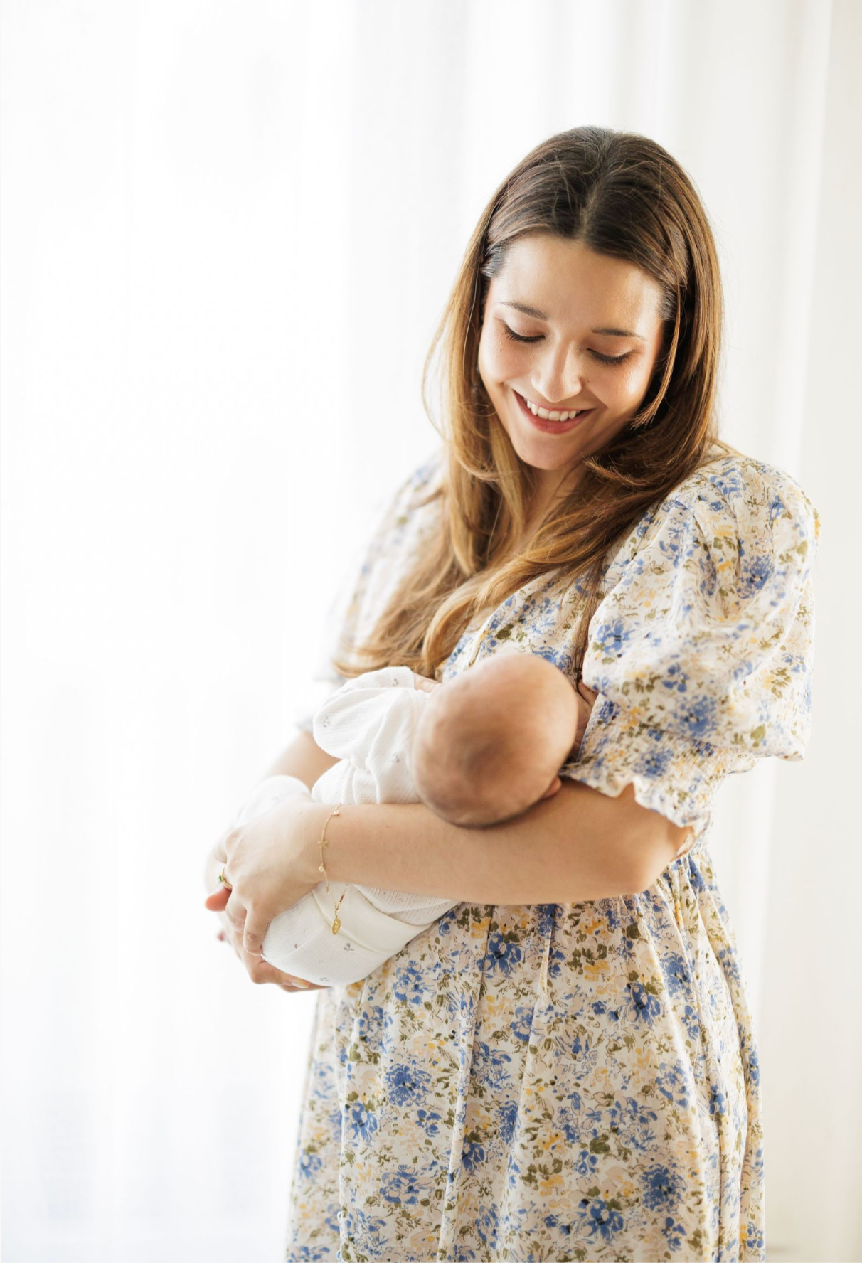Mother in floral dress tenderly holds sleeping newborn near window.