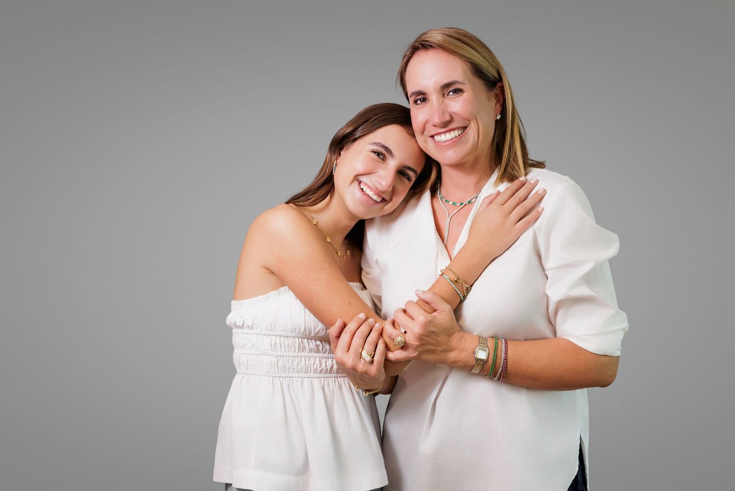 Two women in white outfits embrace warmly while posing against gray background.