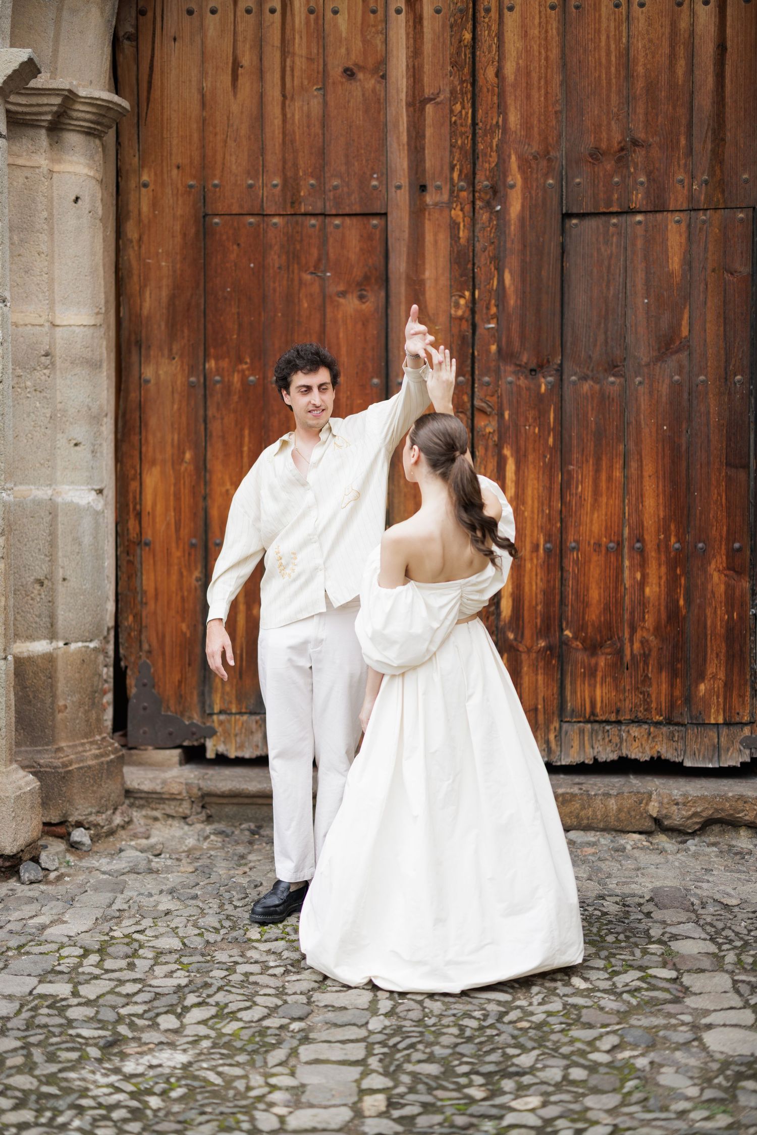 Playful couple in white clothing dancing near rustic wooden doors.