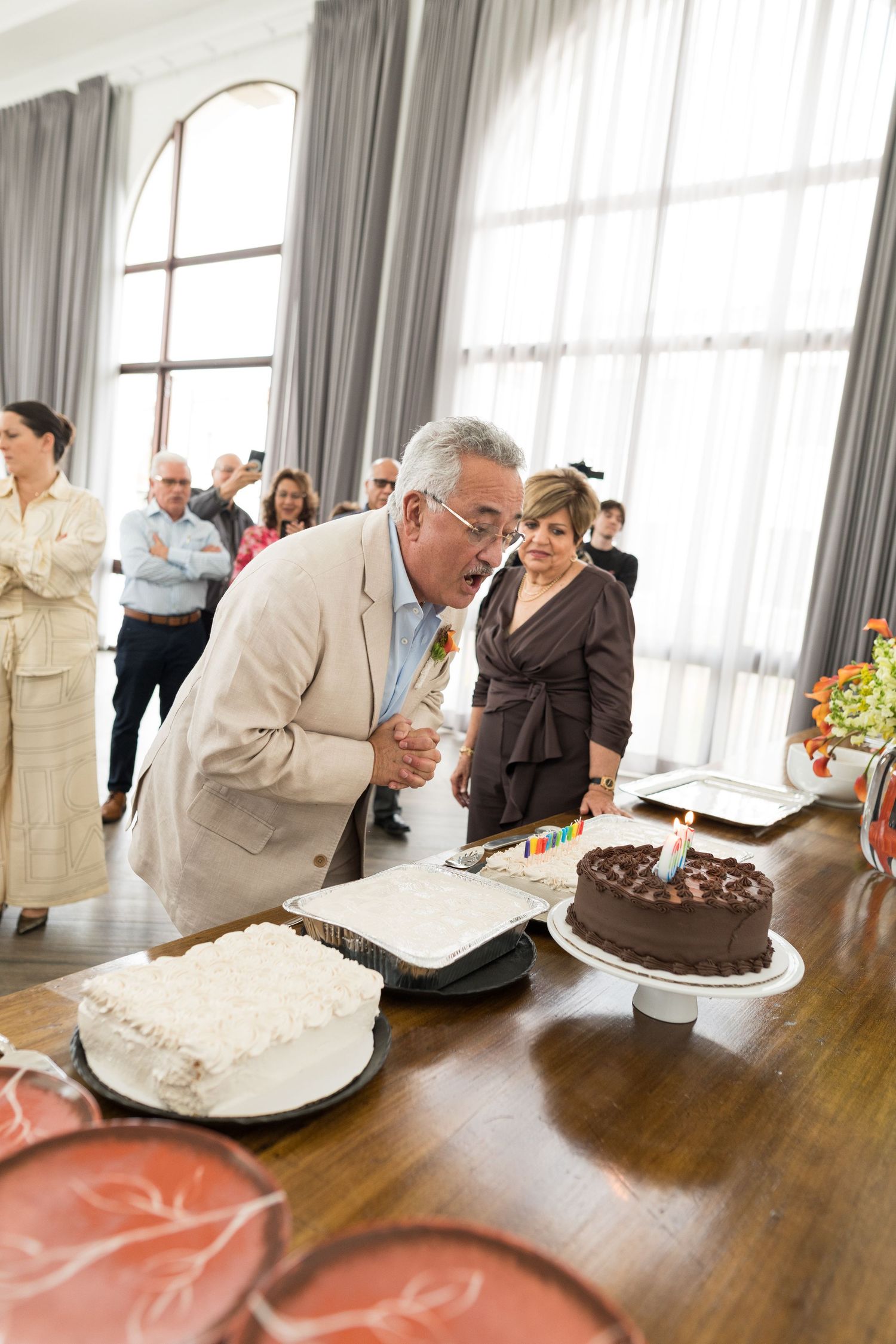 Wedding guests gather around a table with multiple cakes in a bright room with tall windows.