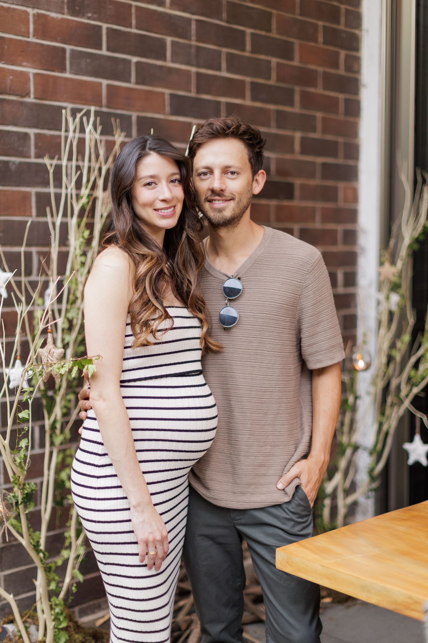 Couple in casual attire posing against brick wall with hanging decorations.