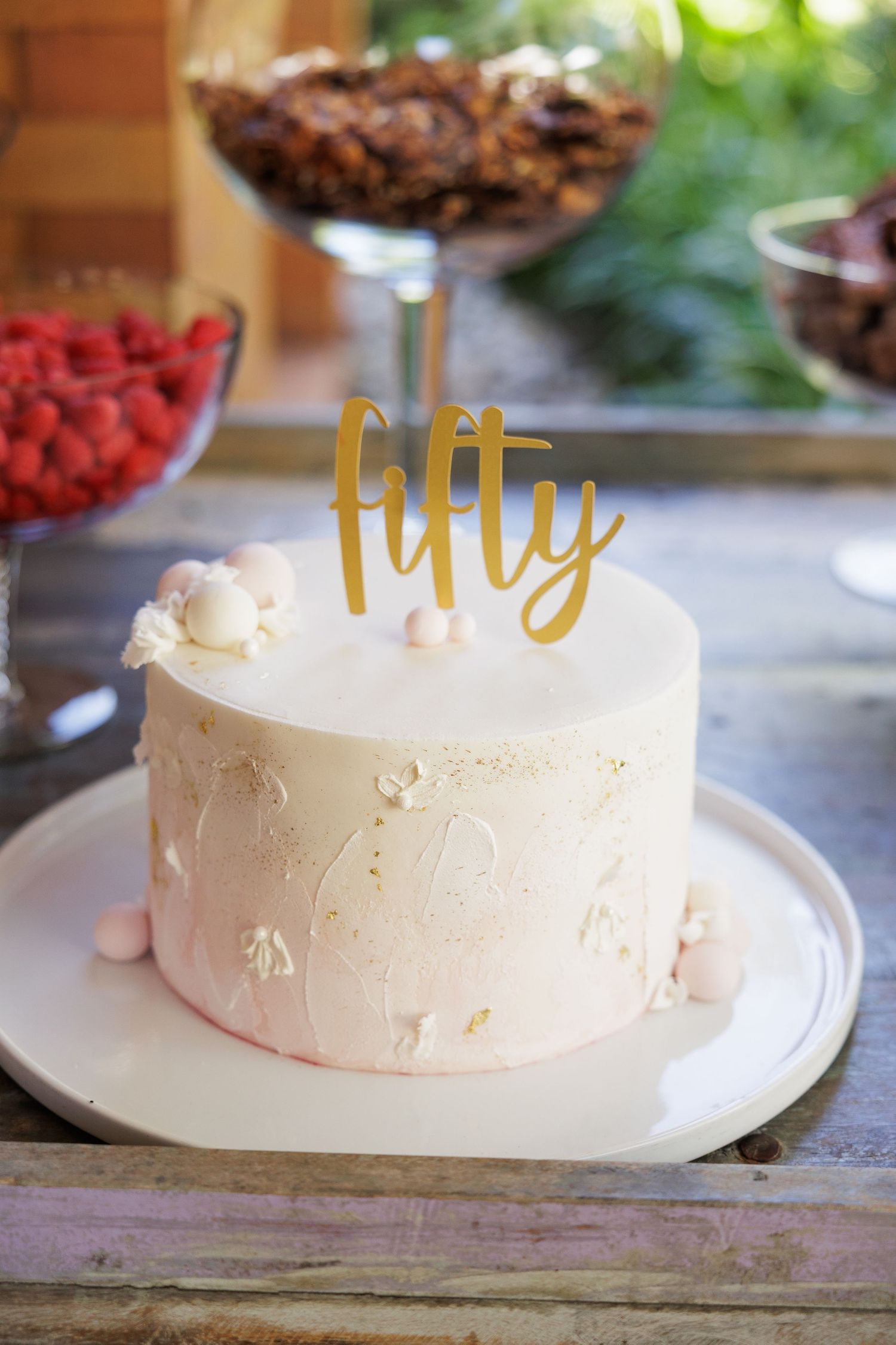 A white frosted cake with gold 'fifty' topper decoration sits on a rustic wooden surface.