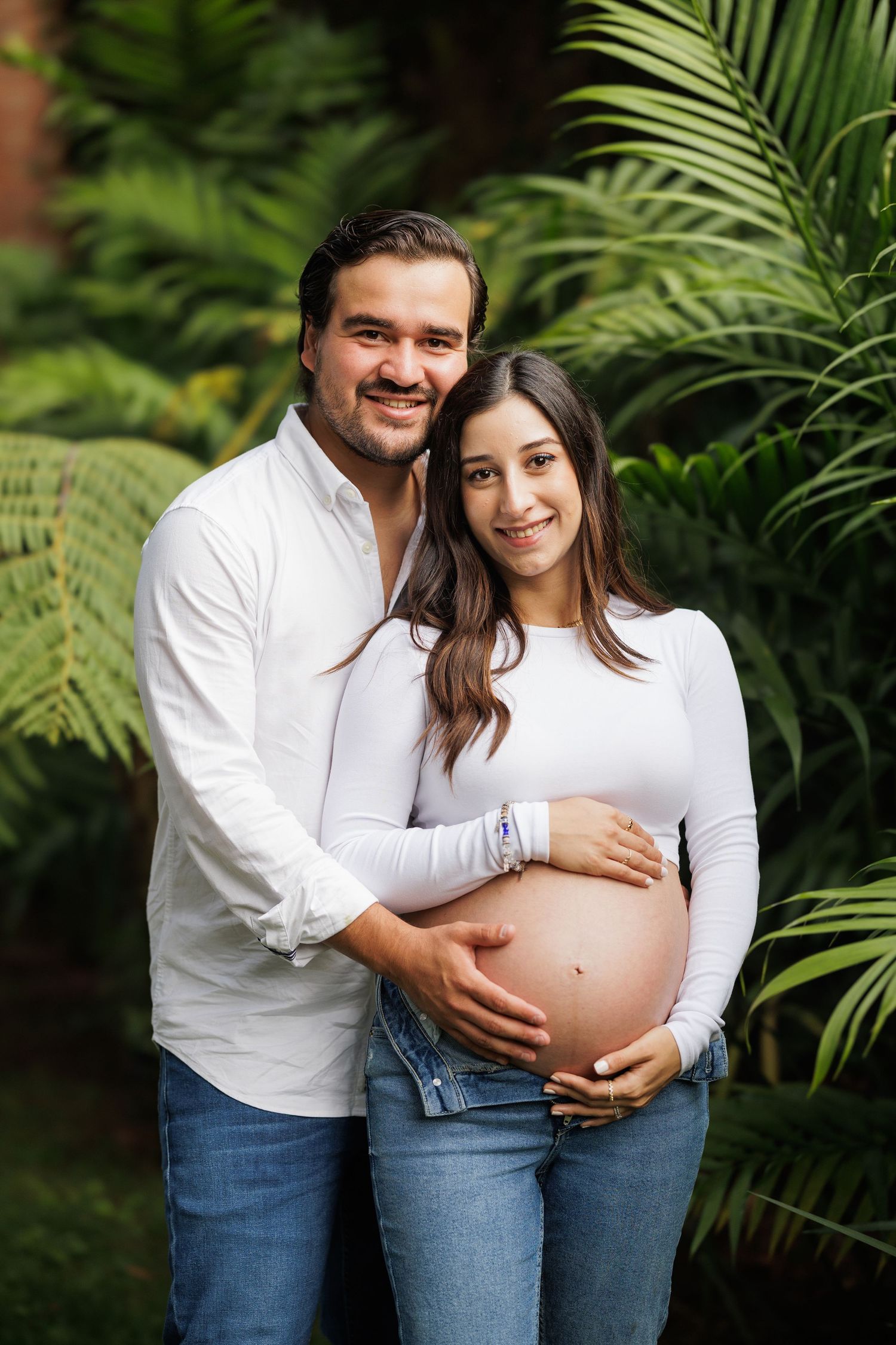 Maternity photoshoot in a lush garden setting with palm fronds creating a tropical backdrop.