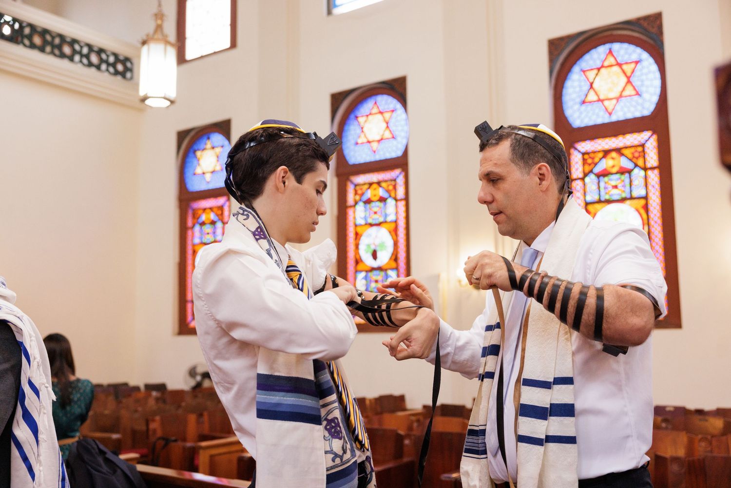 People wearing prayer shawls and kippot gather in a synagogue with stained glass windows during a religious service.