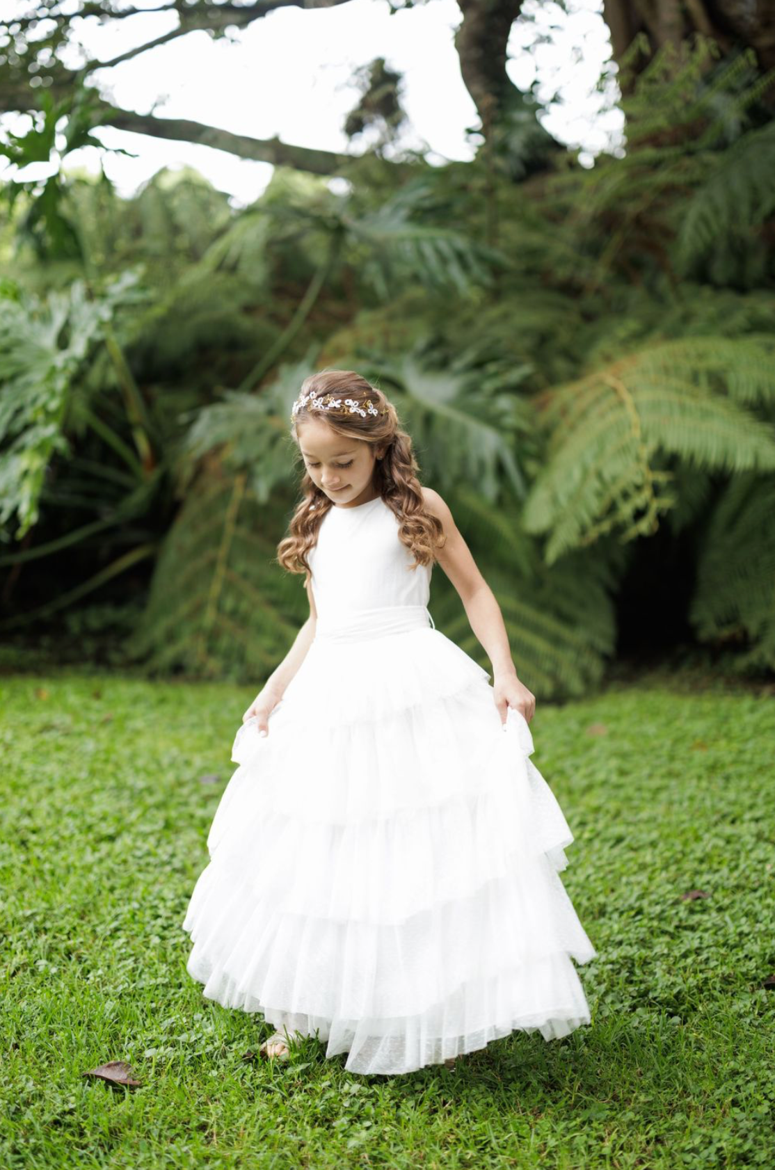Young girl in white layered dress twirls on grass surrounded by lush ferns and greenery.