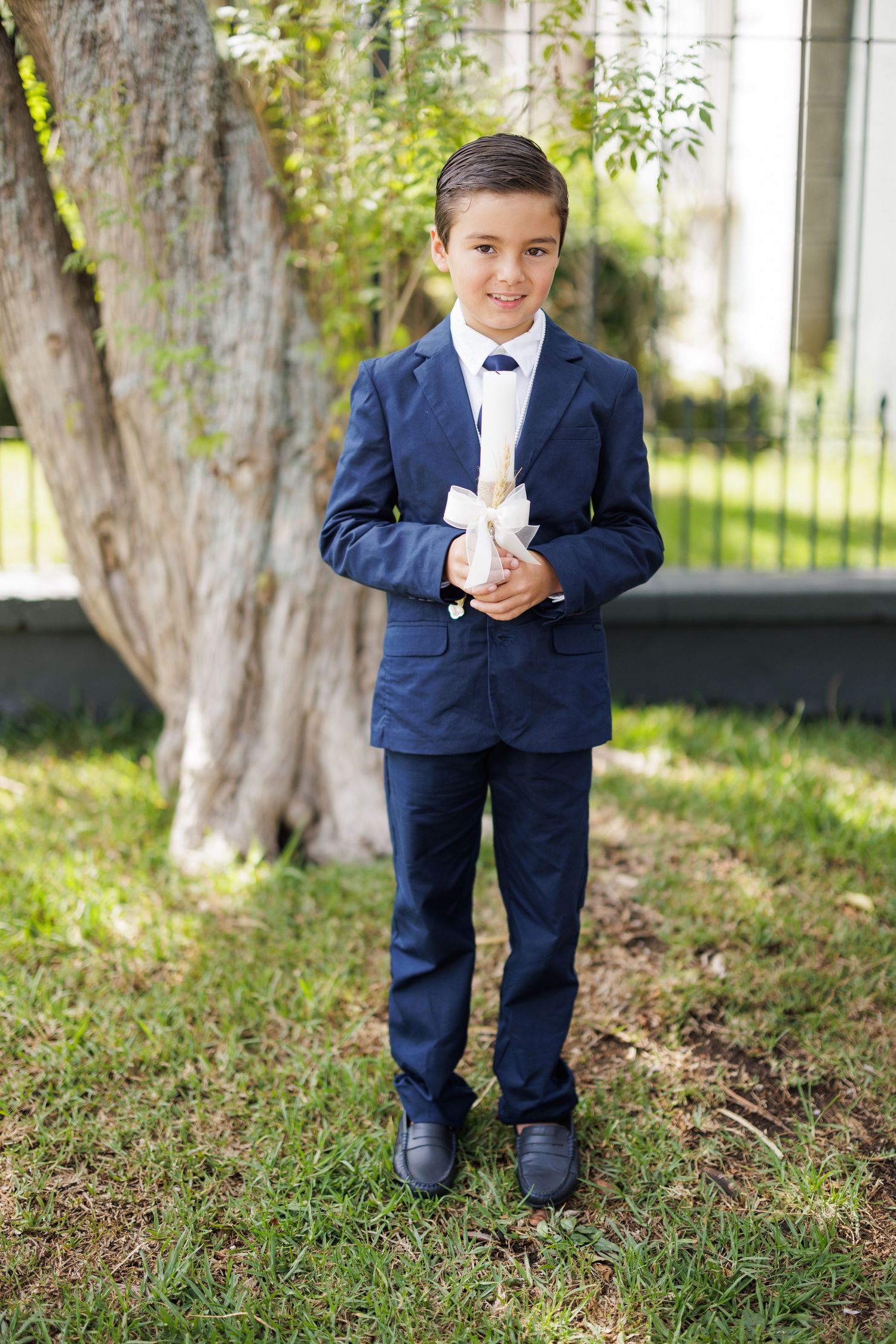 Child in navy suit holds a white candle while standing on grass near trees.