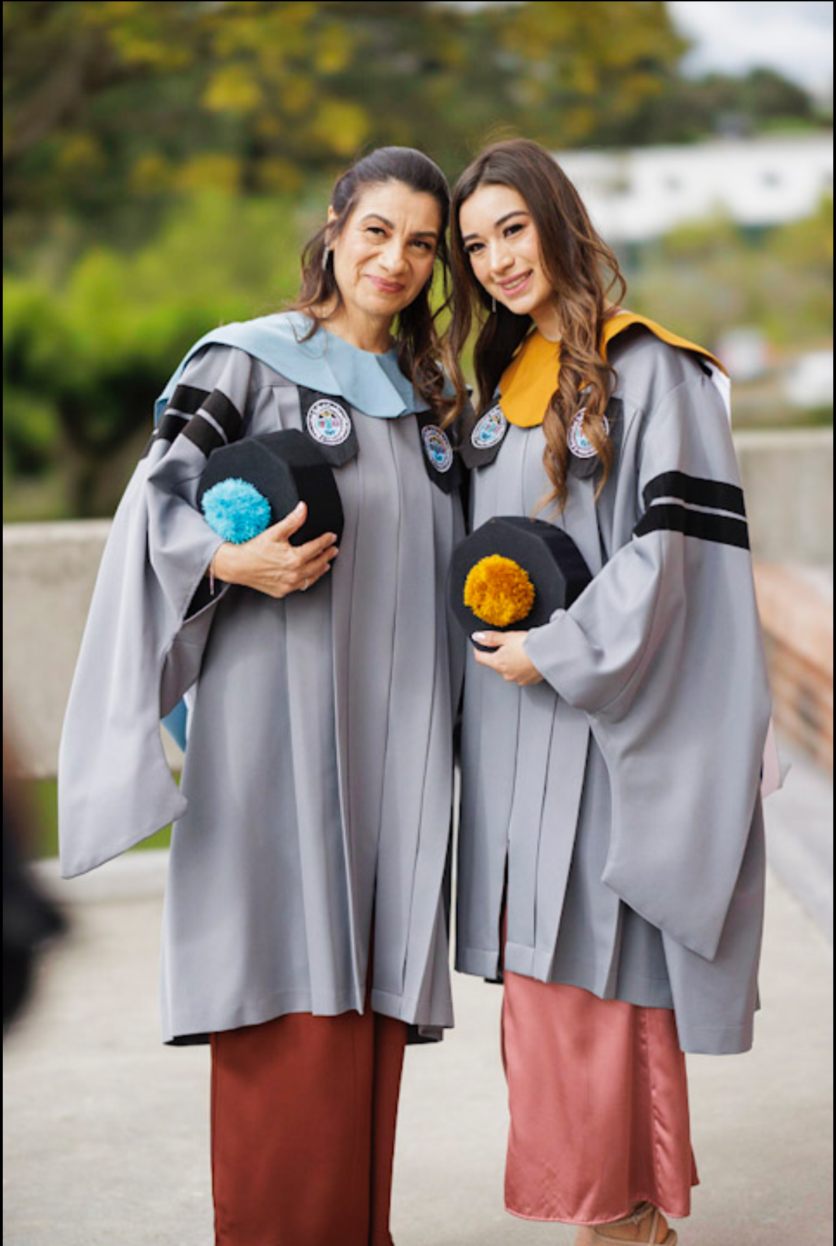 Two graduates in matching grey gowns hold colorful graduation caps during celebration.