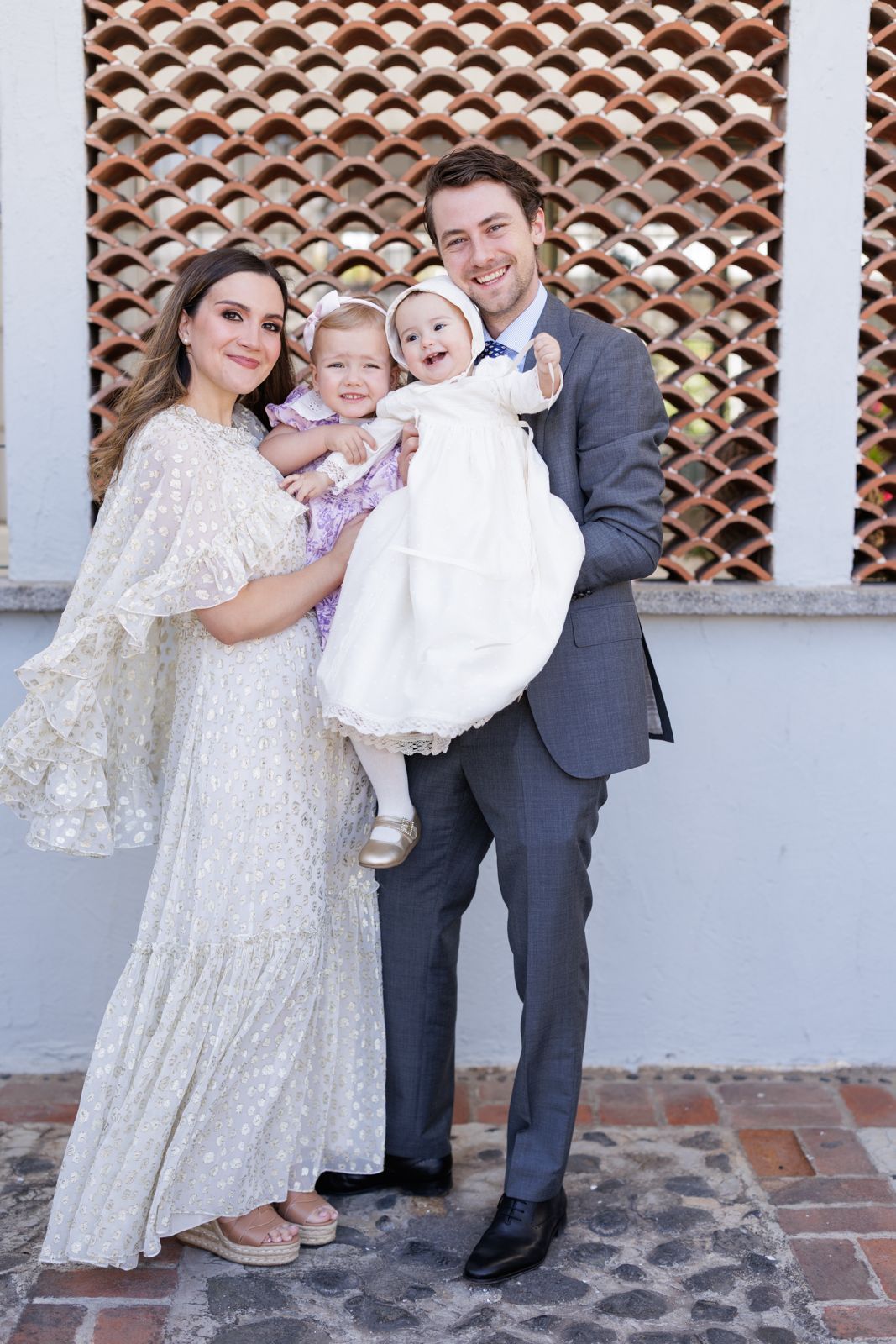 Family in elegant white and gray attire poses together against decorative wall.