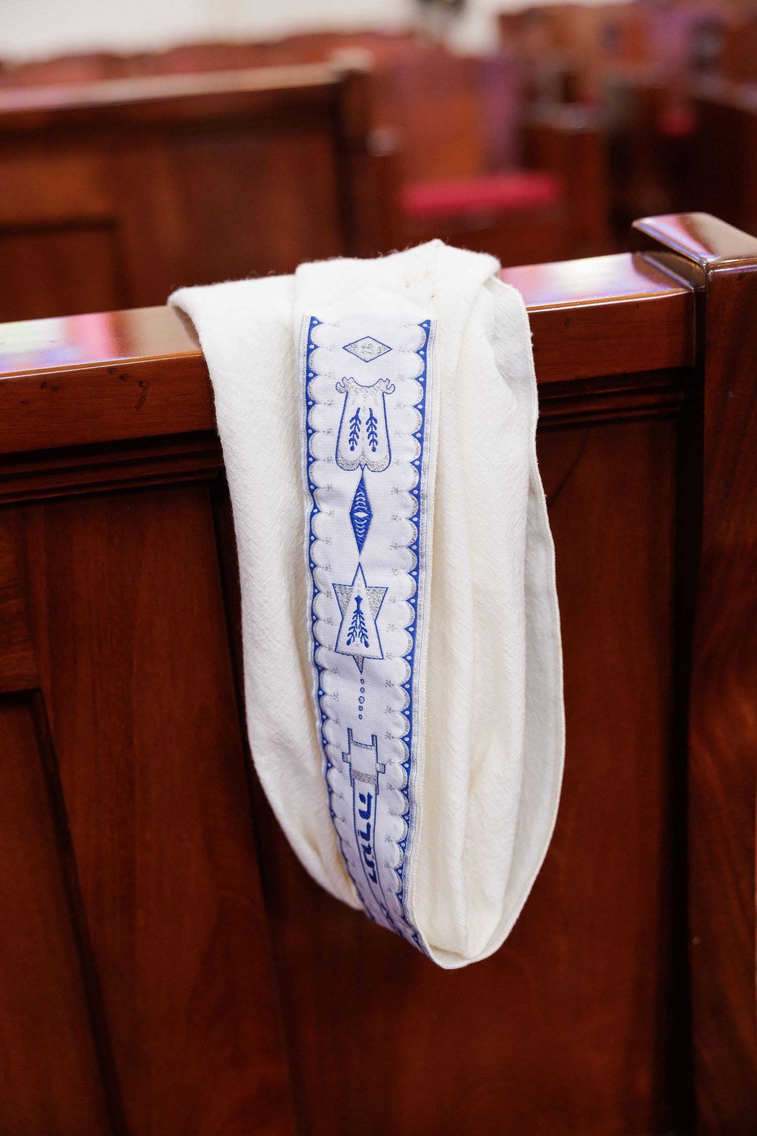 Traditional Jewish prayer shawl with blue embroidery draped over wooden synagogue pew.