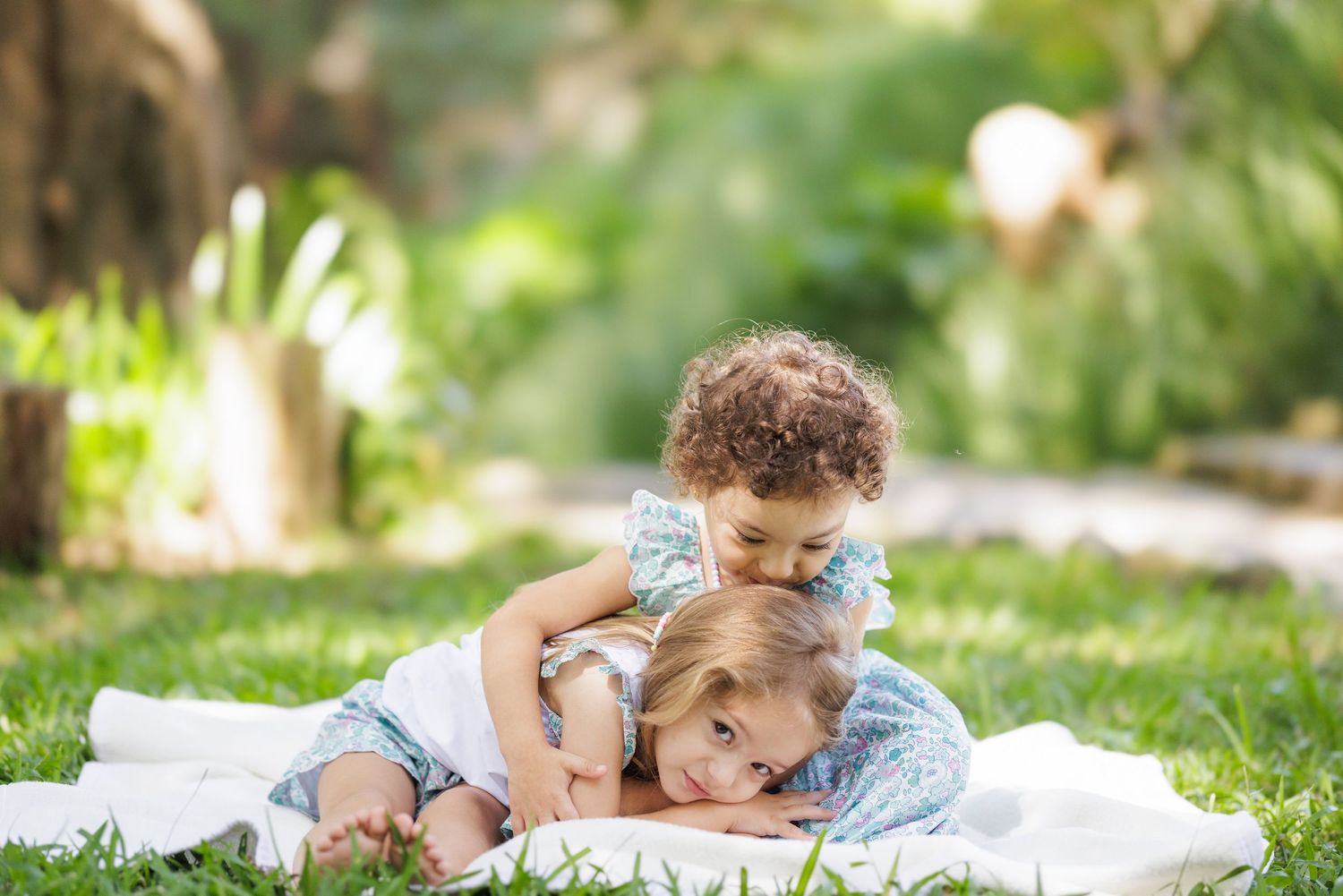 Toddlers in matching outfits play together on blanket in garden setting.