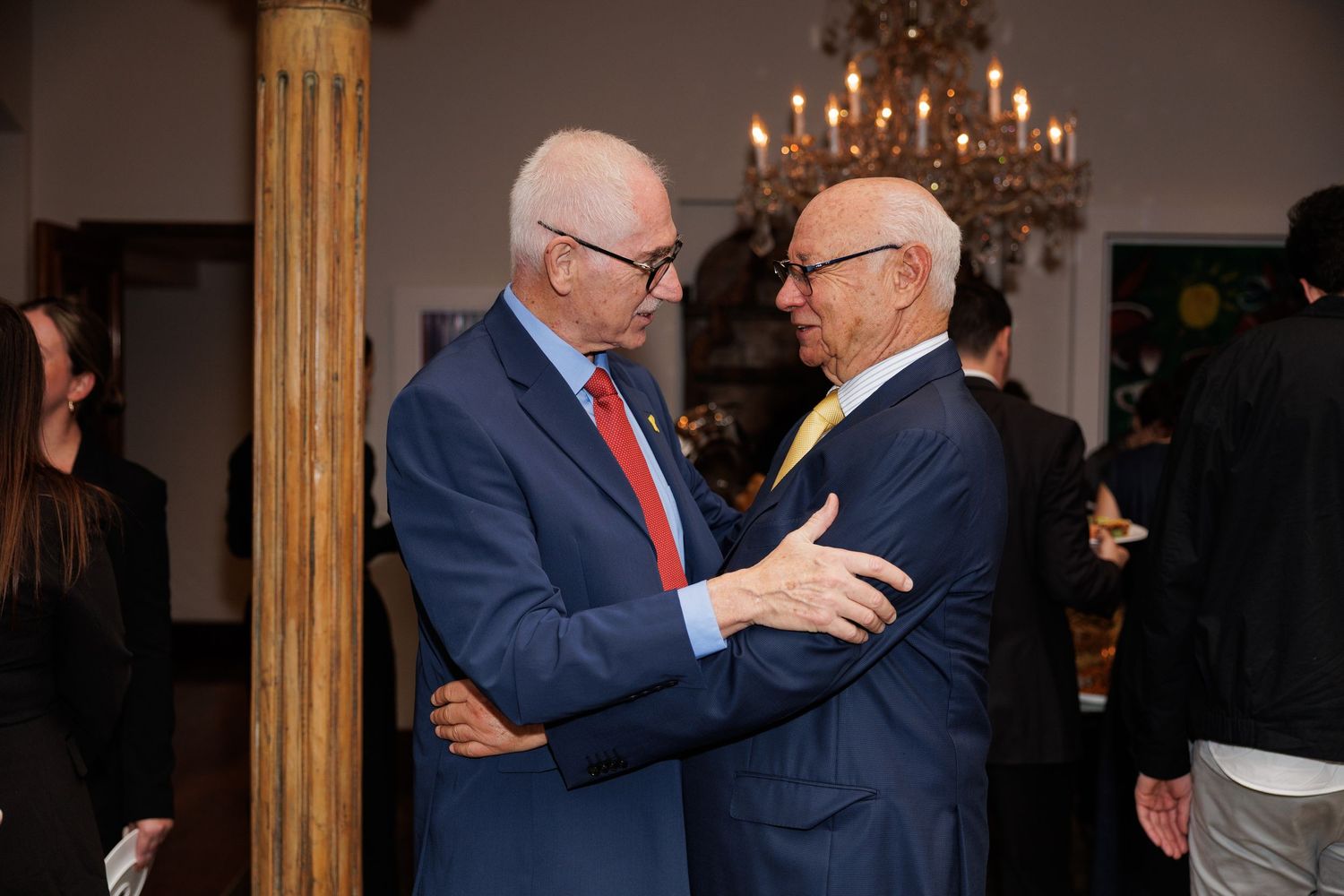 Two men in suits embrace at an elegant indoor event with a chandelier.