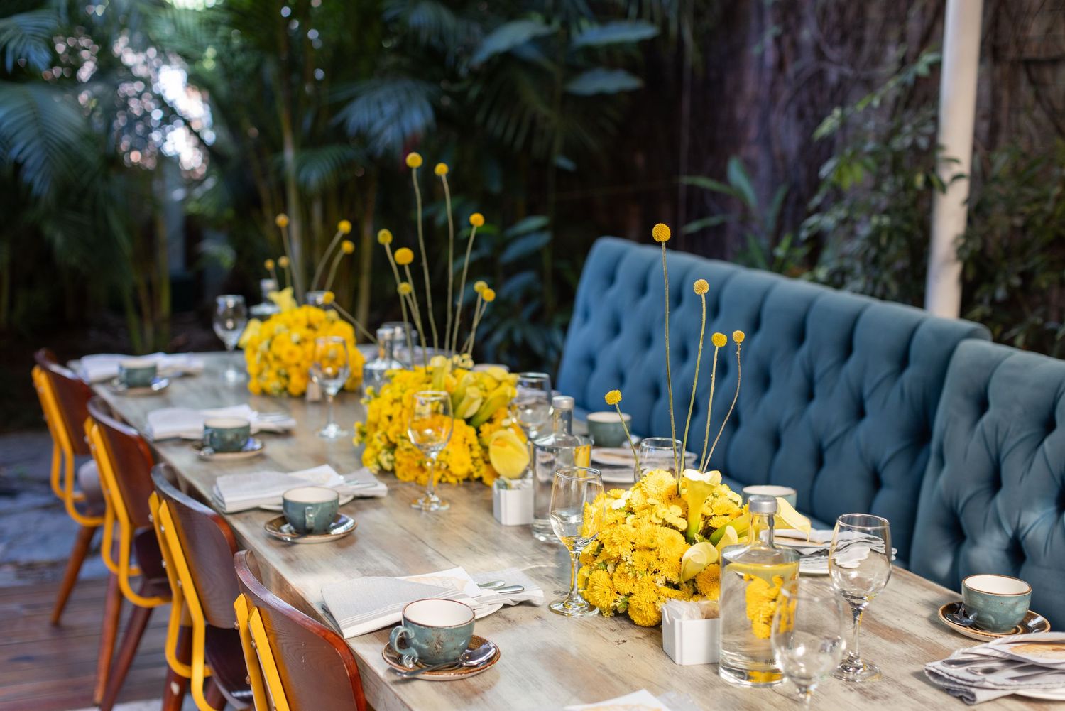 Elegant outdoor wedding reception table with yellow floral centerpieces and tufted blue chairs.