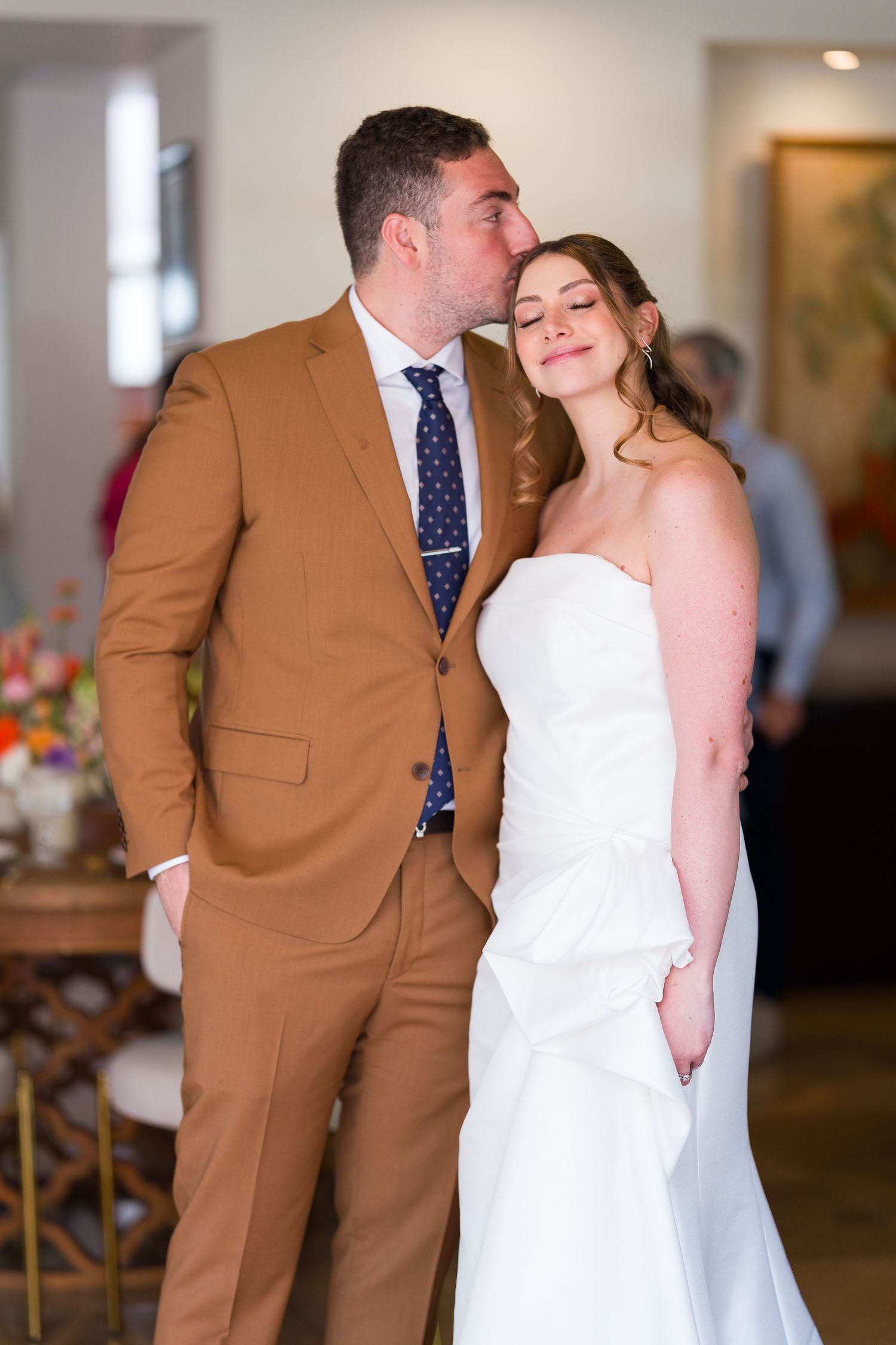 Tender moment between newlyweds in brown suit and white dress indoors.