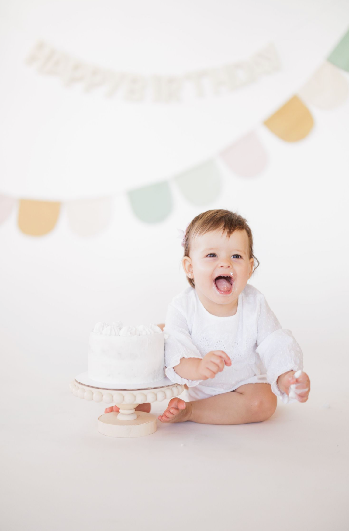 Baby in white outfit sits smiling with birthday cake under pastel party decorations.