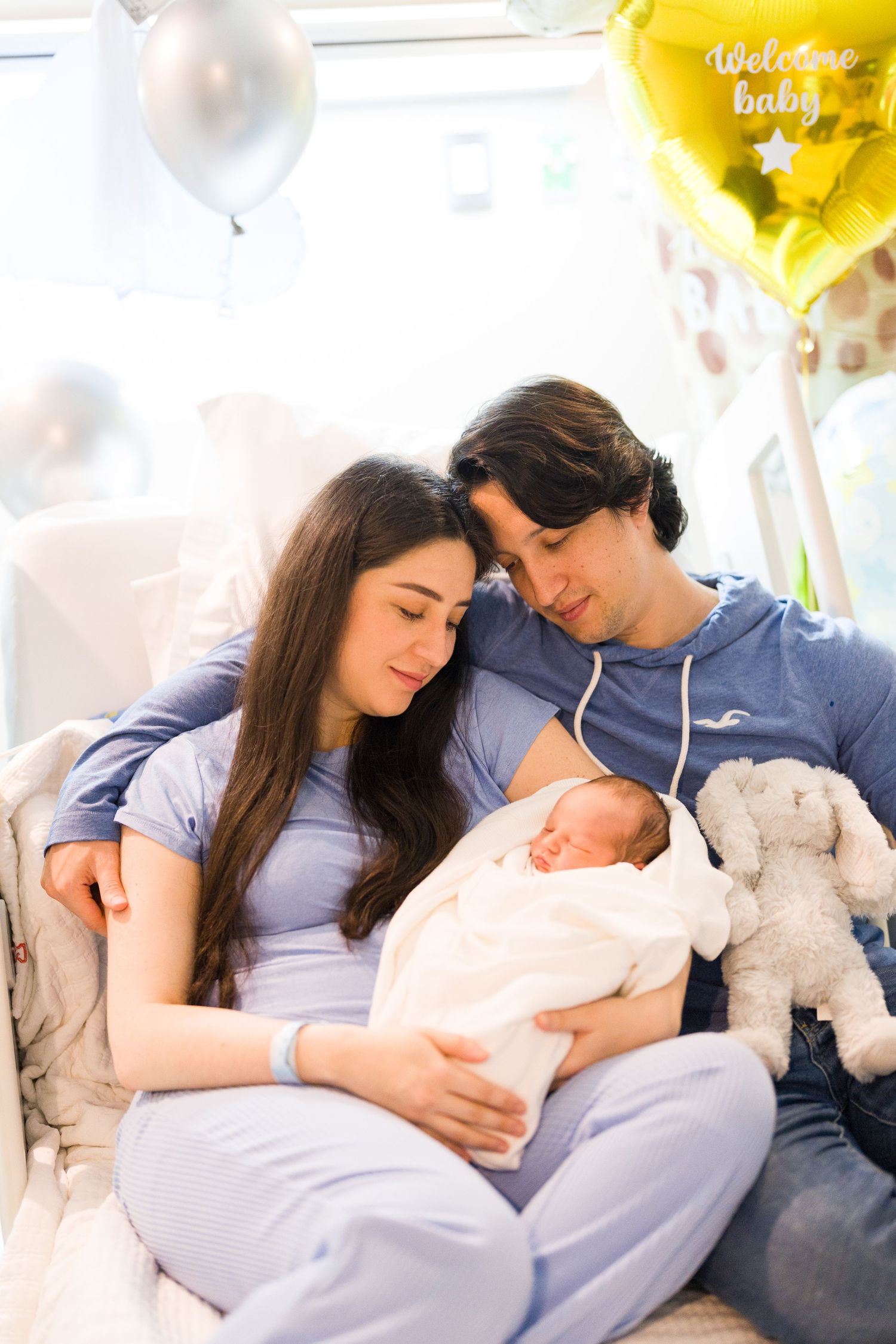 New parents cuddle with their newborn baby on a couch surrounded by yellow balloons and stuffed animals.