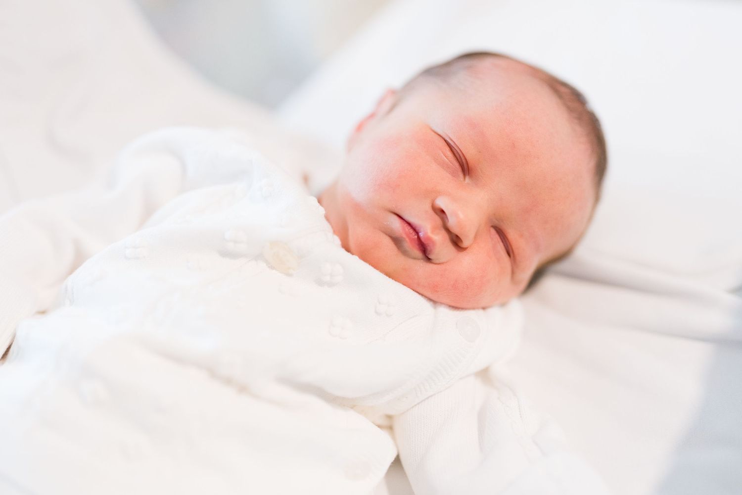 Peaceful sleeping newborn wearing all white against light background.