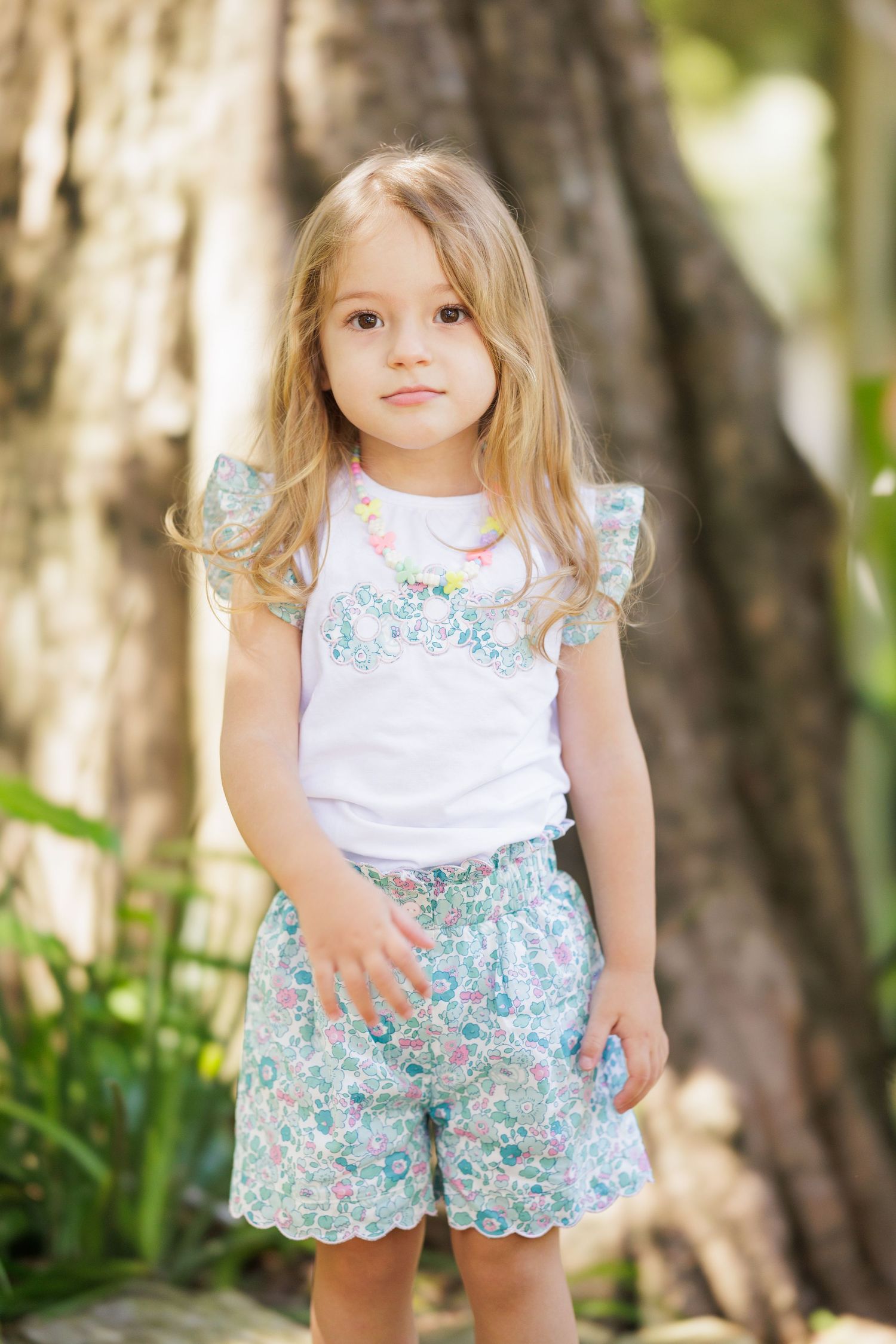 Child wearing floral shorts and white top poses in natural outdoor setting.