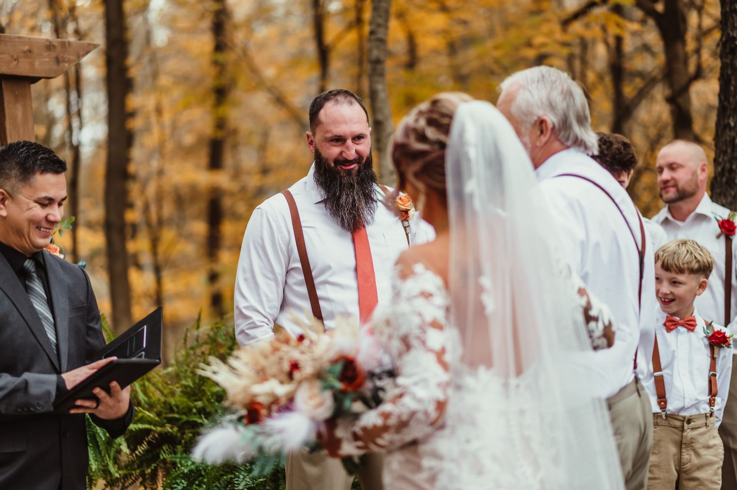 Groom smiling during outdoor fall wedding ceremony with officiant, bride, and guests among autumn foliage.