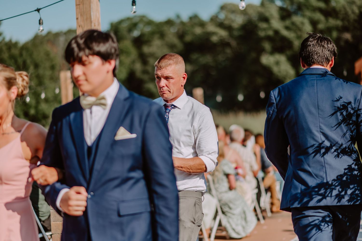 Groomsmen in navy suits gather near wooden ceremony arch at an outdoor wedding venue.