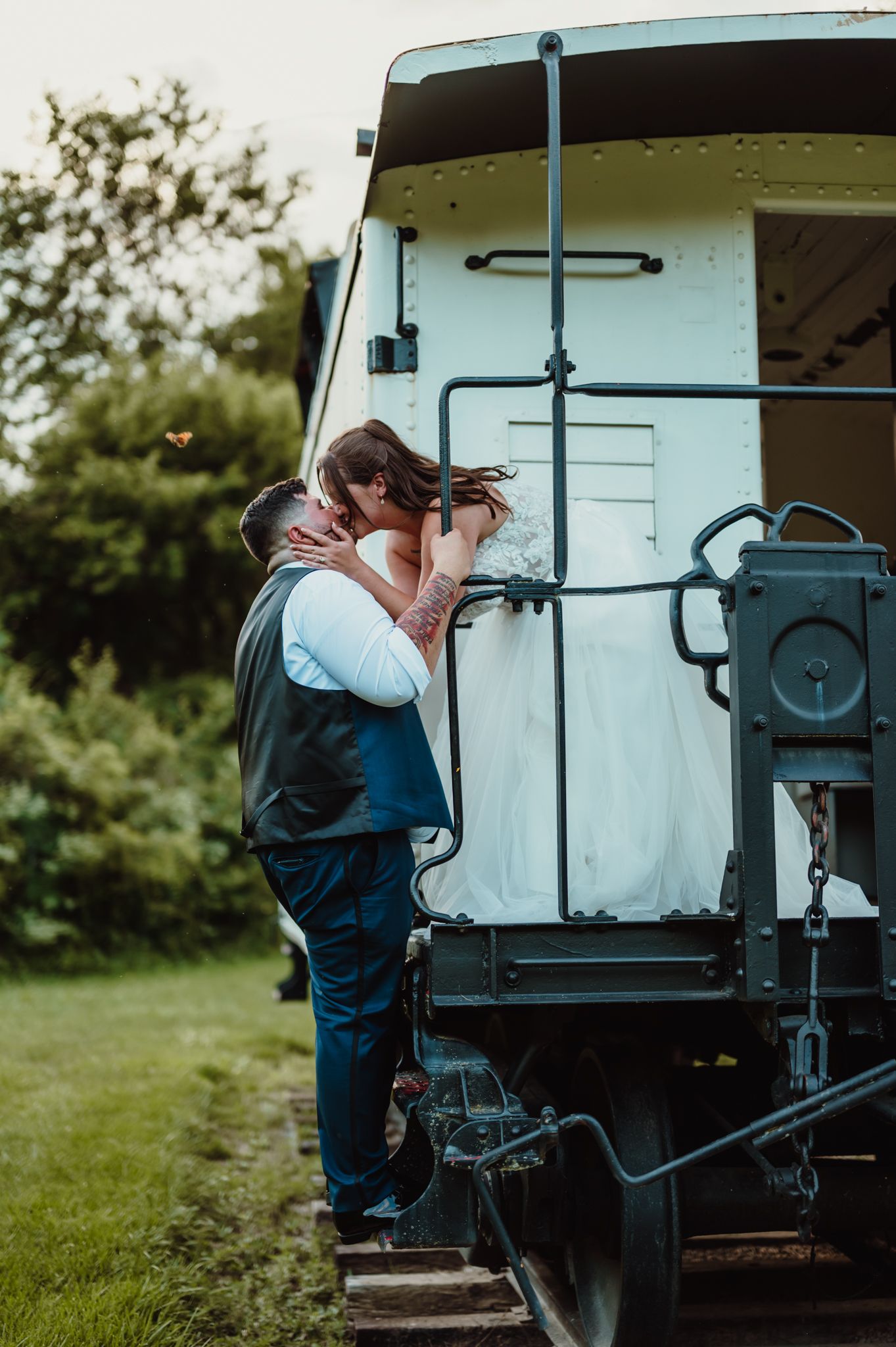 A bride and groom share a romantic kiss on the back of a vintage train car surrounded by lush greenery.