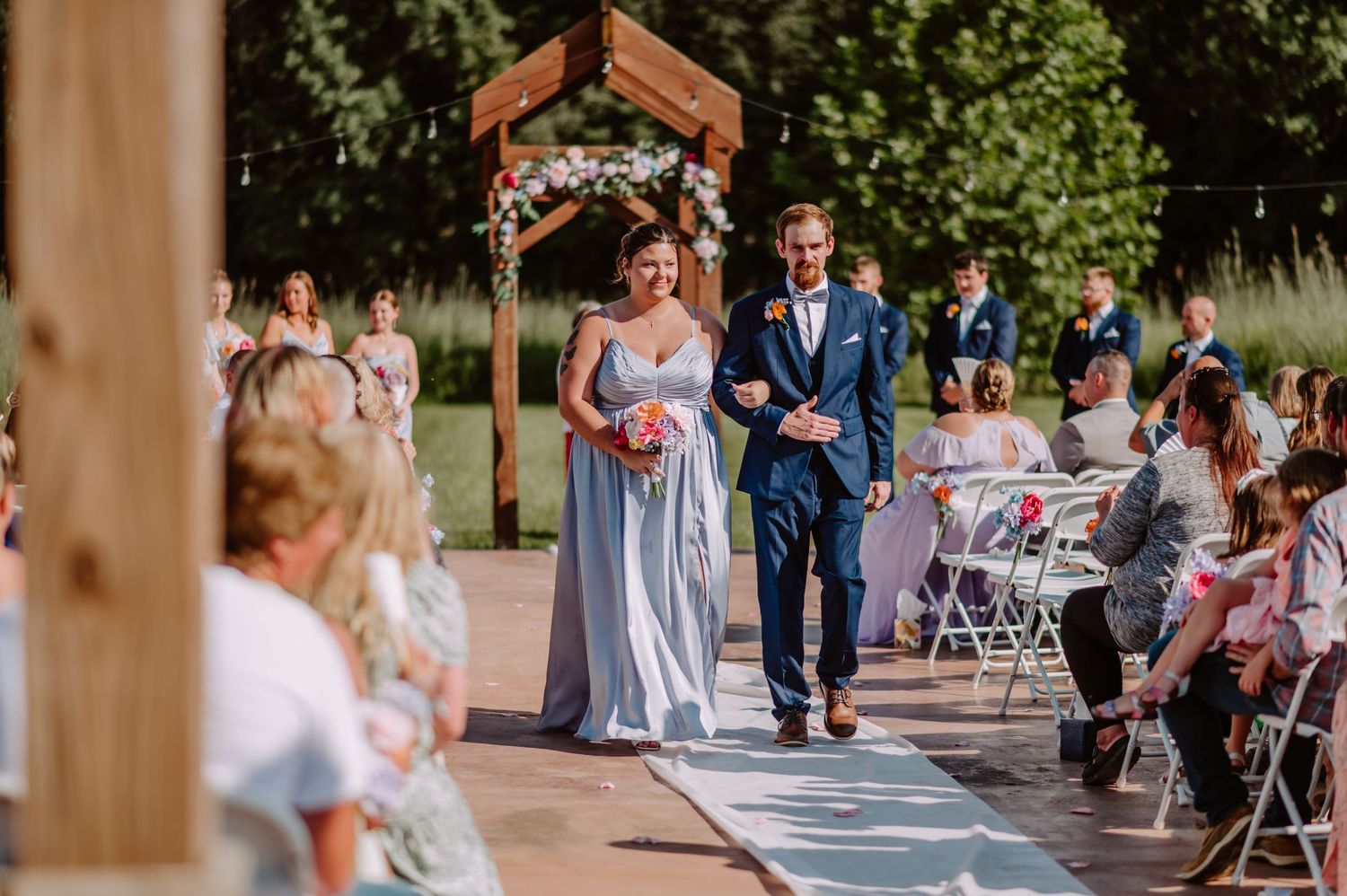 Bridesmaid in light blue gown walks arm-in-arm with groomsman down sunlit outdoor wedding aisle.