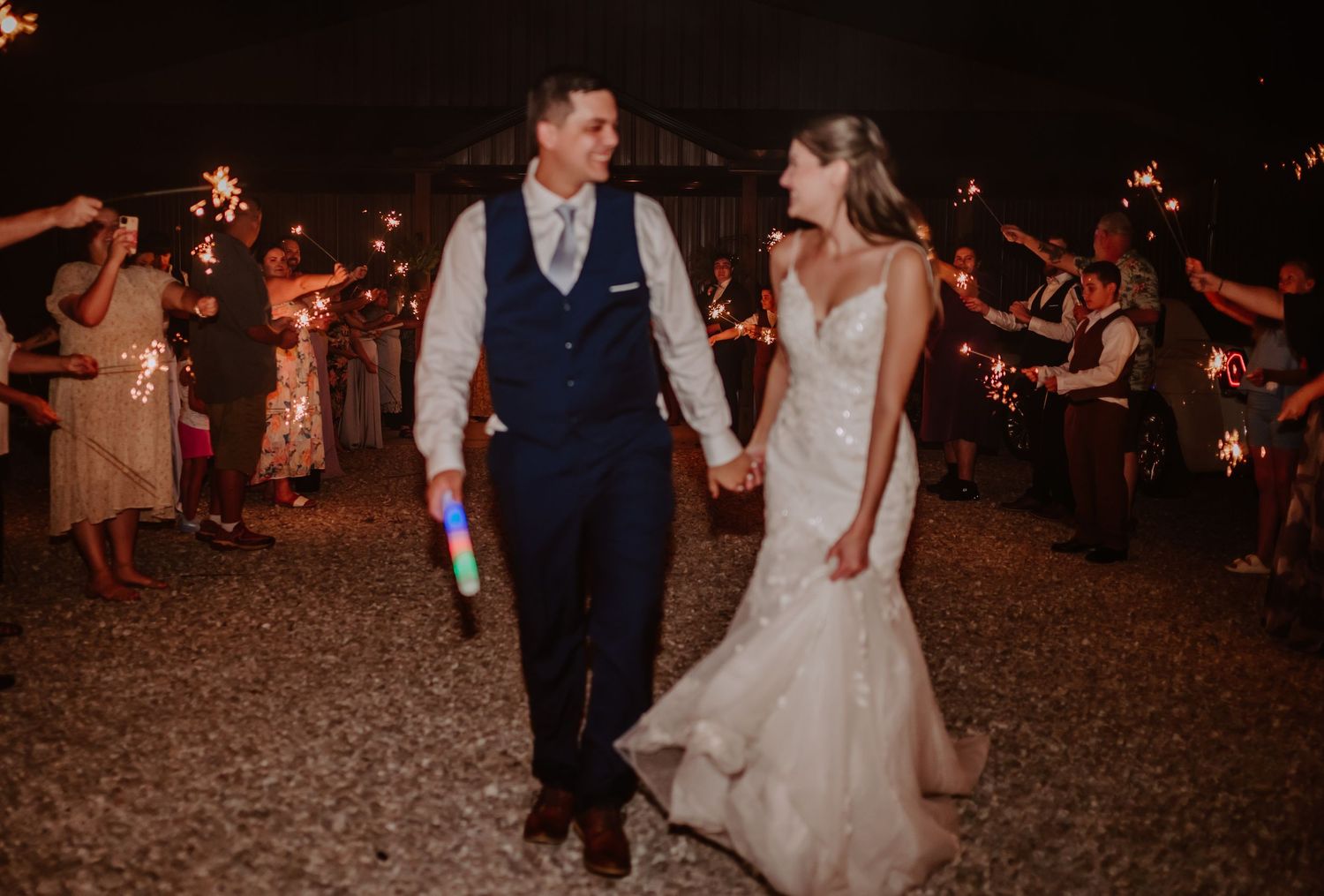Newlyweds hold hands walking through sparkler sendoff at romantic outdoor wedding reception at night.