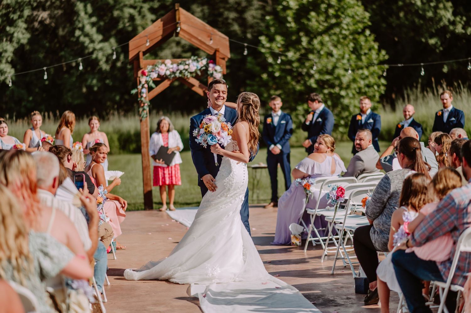Groom lifts bride during recessional at outdoor wedding ceremony with floral wooden arch.