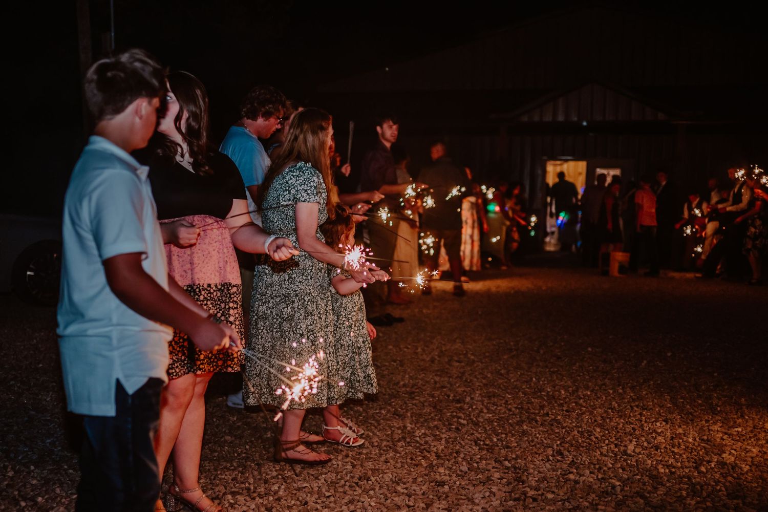 Wedding guests hold lit sparklers in line during nighttime outdoor wedding sendoff celebration.