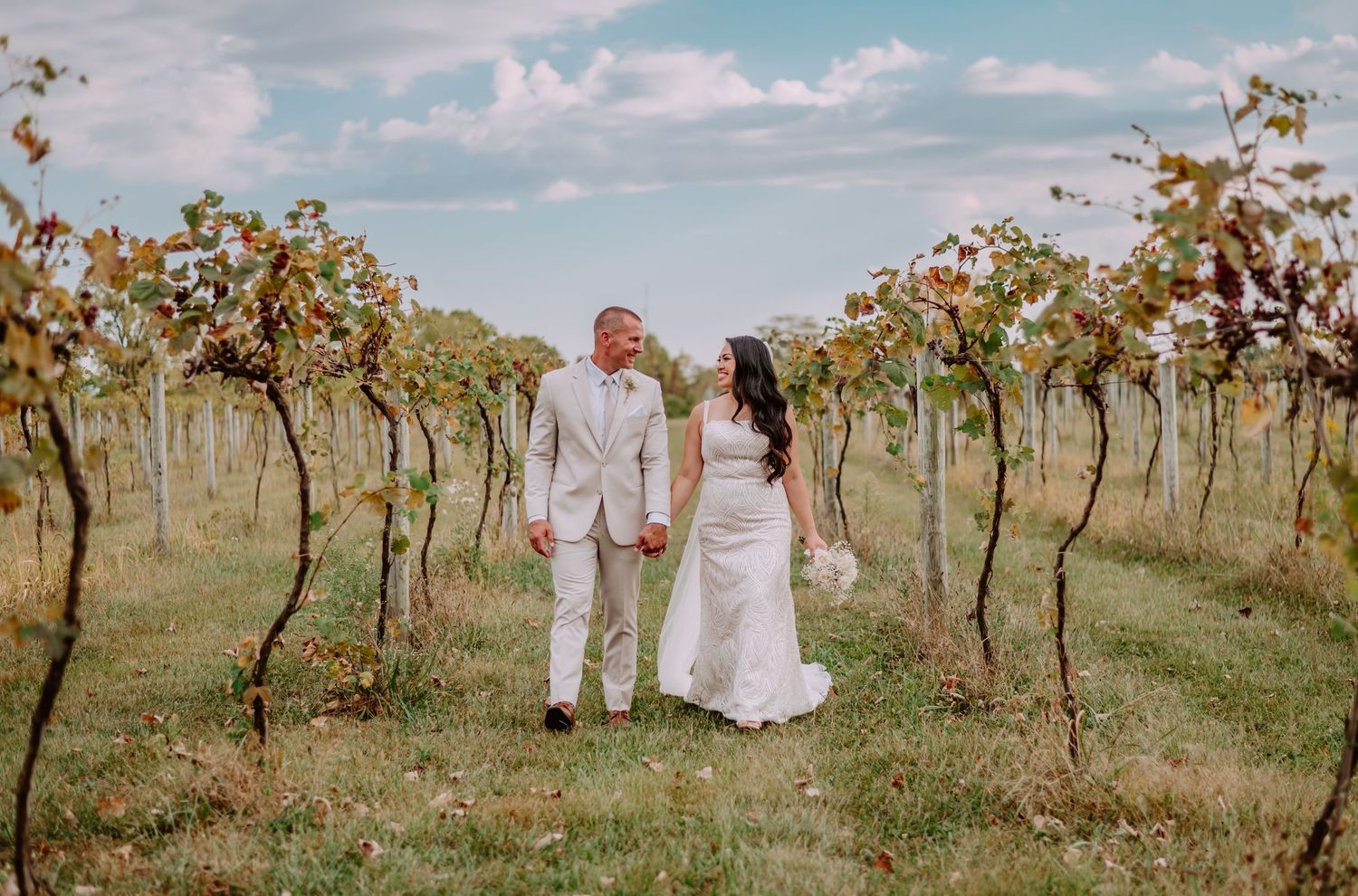 Bride and groom holding hands and smiling in a lush vineyard on their wedding day under a cloudy sky.
