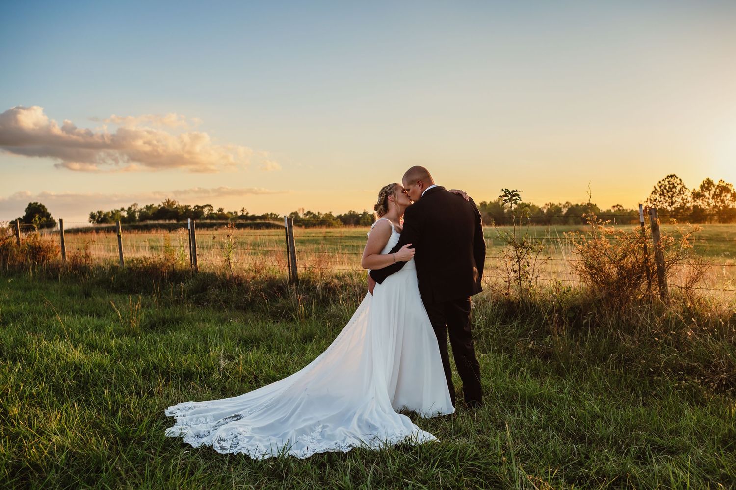 Bride in flowing white gown and groom embracing at sunset beside rustic fence in field.