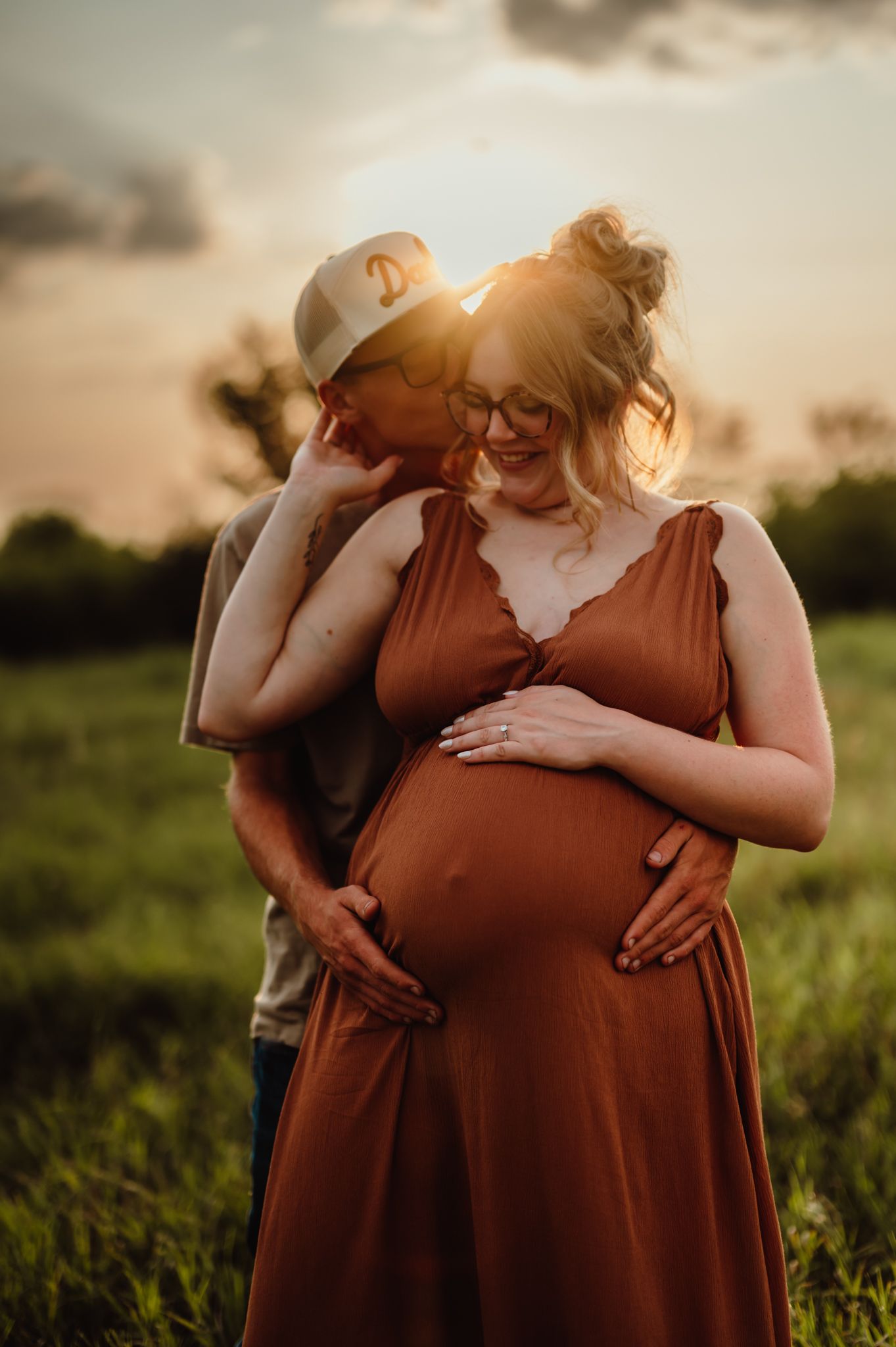 A couple shares a tender sunset maternity moment in a green field, the woman in a rust dress cradling her baby bump.