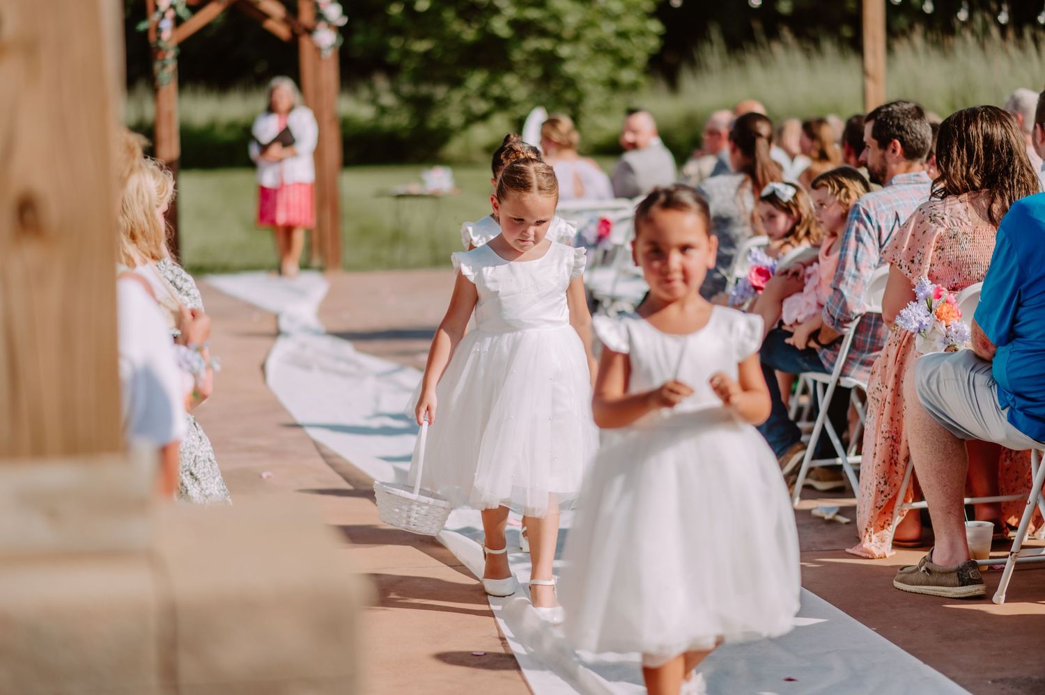 Two young flower girls in white dresses walk down the wedding aisle during an outdoor ceremony.