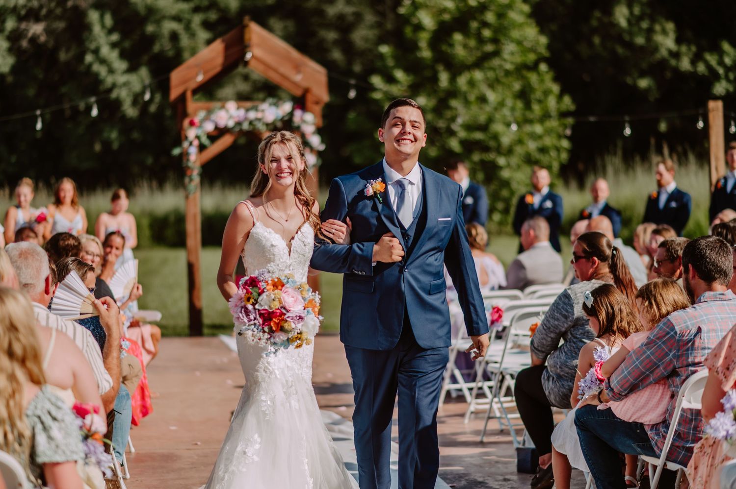 Smiling newlyweds walk arm-in-arm down outdoor wedding aisle past cheering guests under string lights.