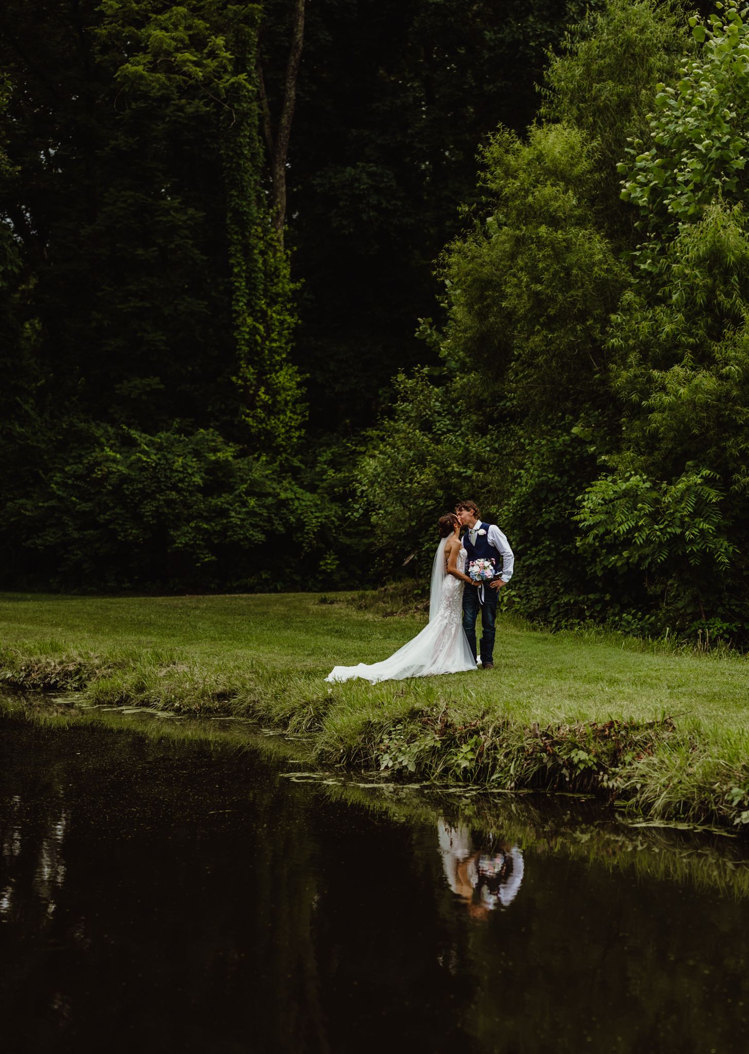 A bride and groom share a romantic kiss beside a reflective pond surrounded by lush green trees on their wedding day.
