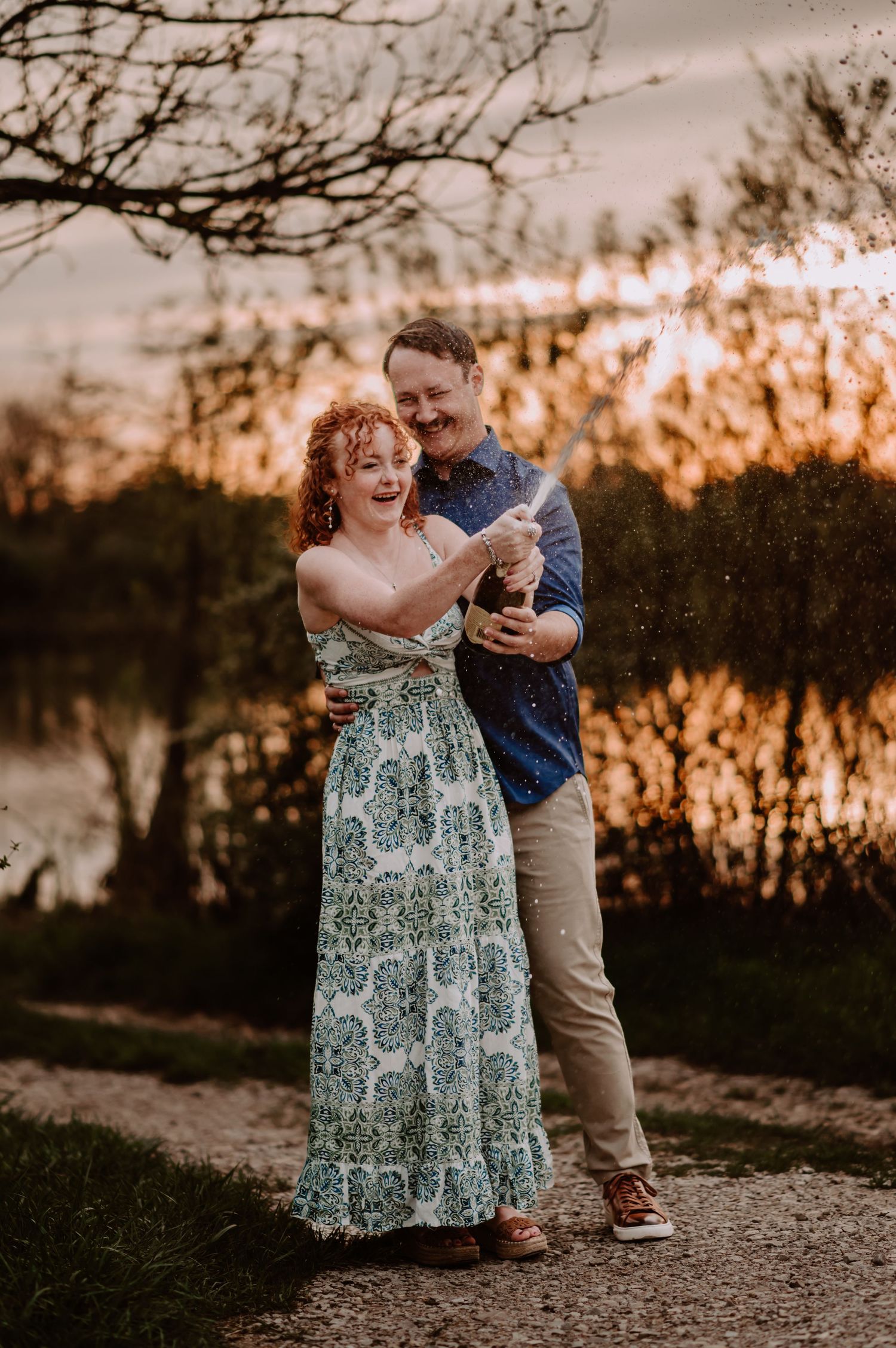 Couple laughing and popping champagne bottle outdoors at golden sunset near a lake, woman in floral dress, man in blue shirt.