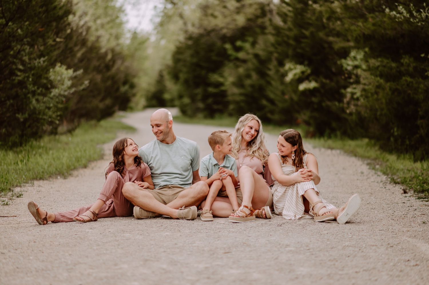Family of six sitting together on a dirt path surrounded by lush green trees, smiling and enjoying a candid outdoor moment.