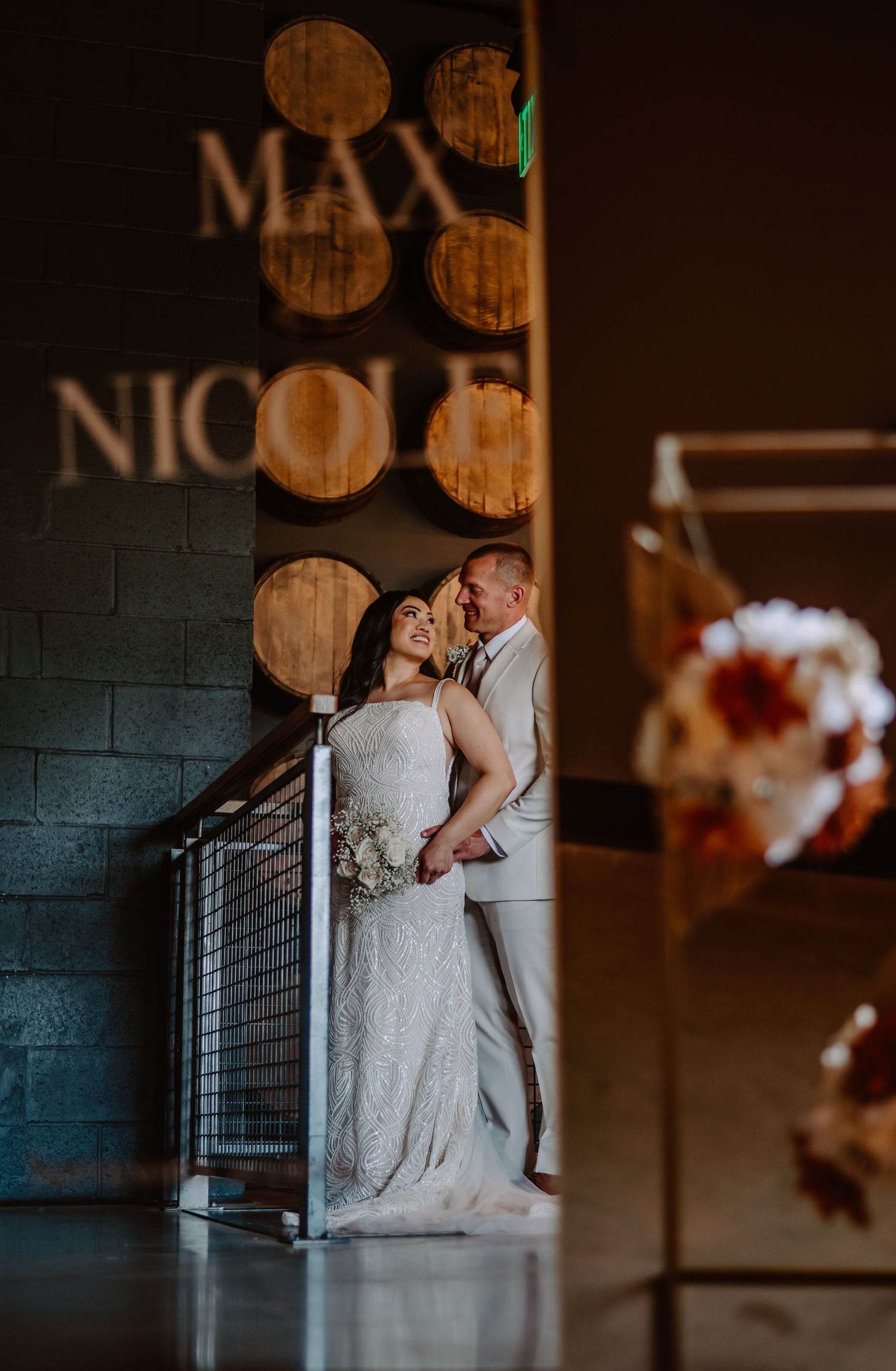 Bride and groom share a romantic moment at a winery wedding venue with barrel decor and their names MAX and NICOLE illuminated.