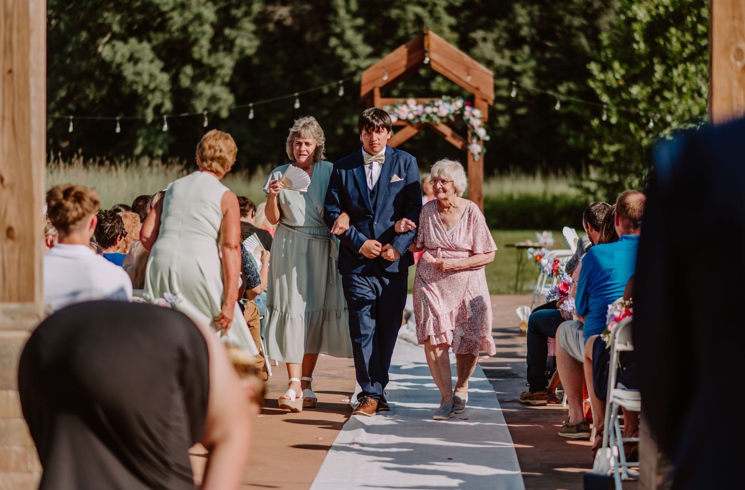 Groom escorts two women down the wedding aisle at an outdoor ceremony with wooden arch.