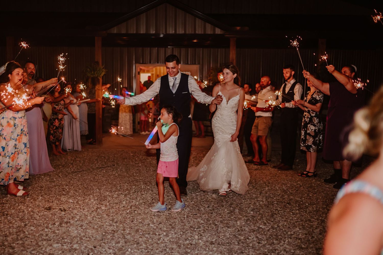 Bride and groom walk through sparkler tunnel with child holding glow stick at outdoor wedding reception at night.