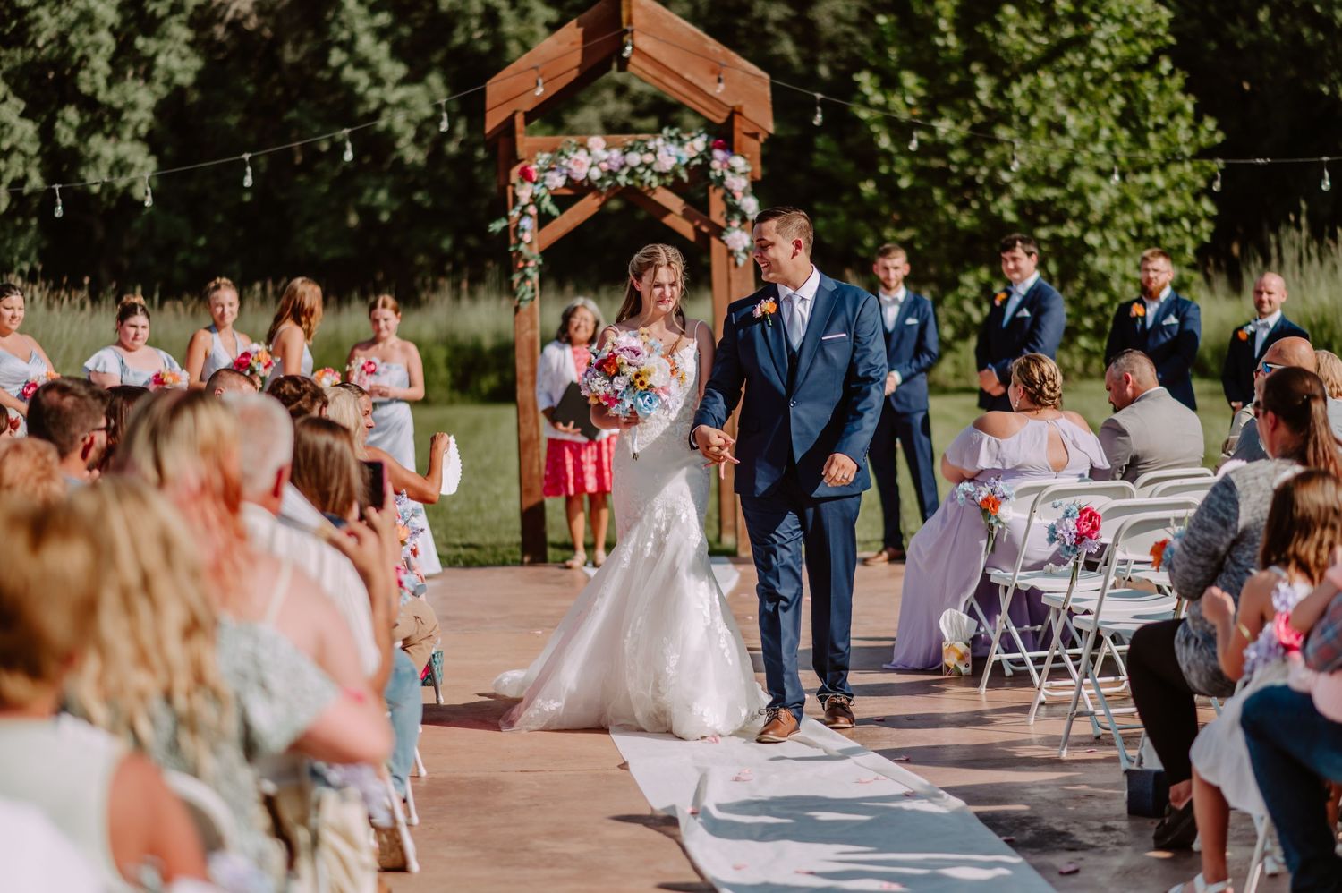 Newlyweds walk hand in hand down aisle at outdoor wedding ceremony surrounded by guests.