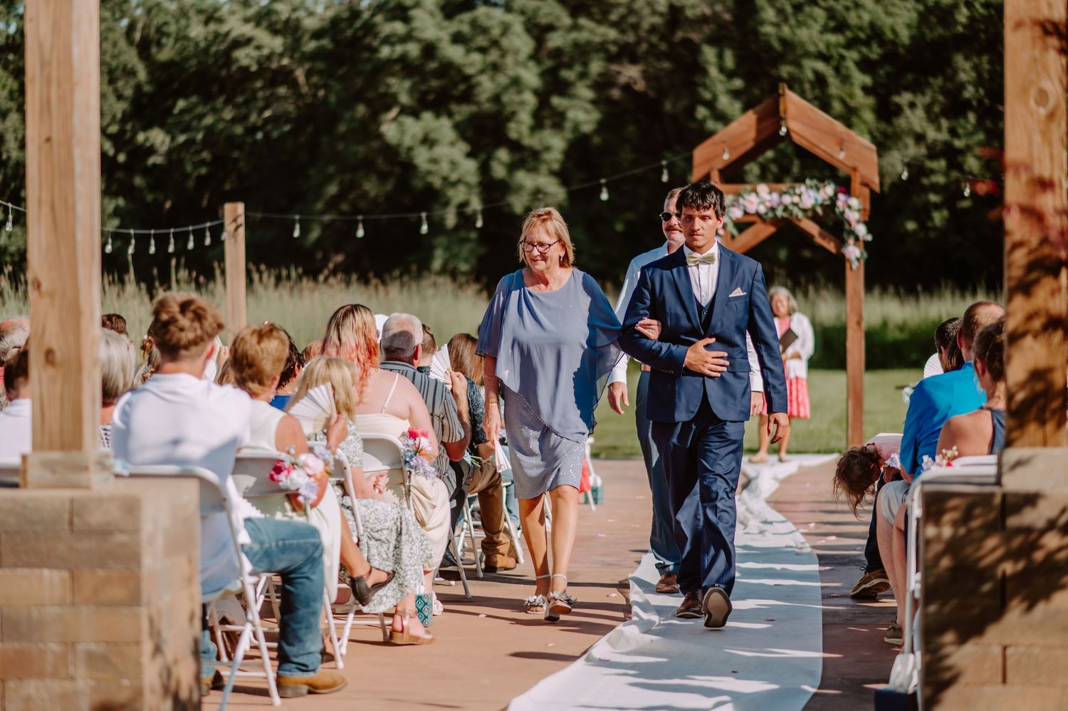 Groom in navy suit walks down outdoor wedding aisle escorted by woman in blue dress past seated guests.
