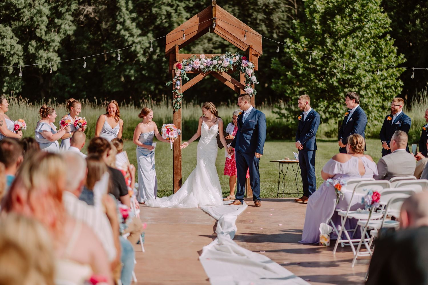 Bride and groom exchange vows under wooden arch with pink flowers at outdoor wedding ceremony.