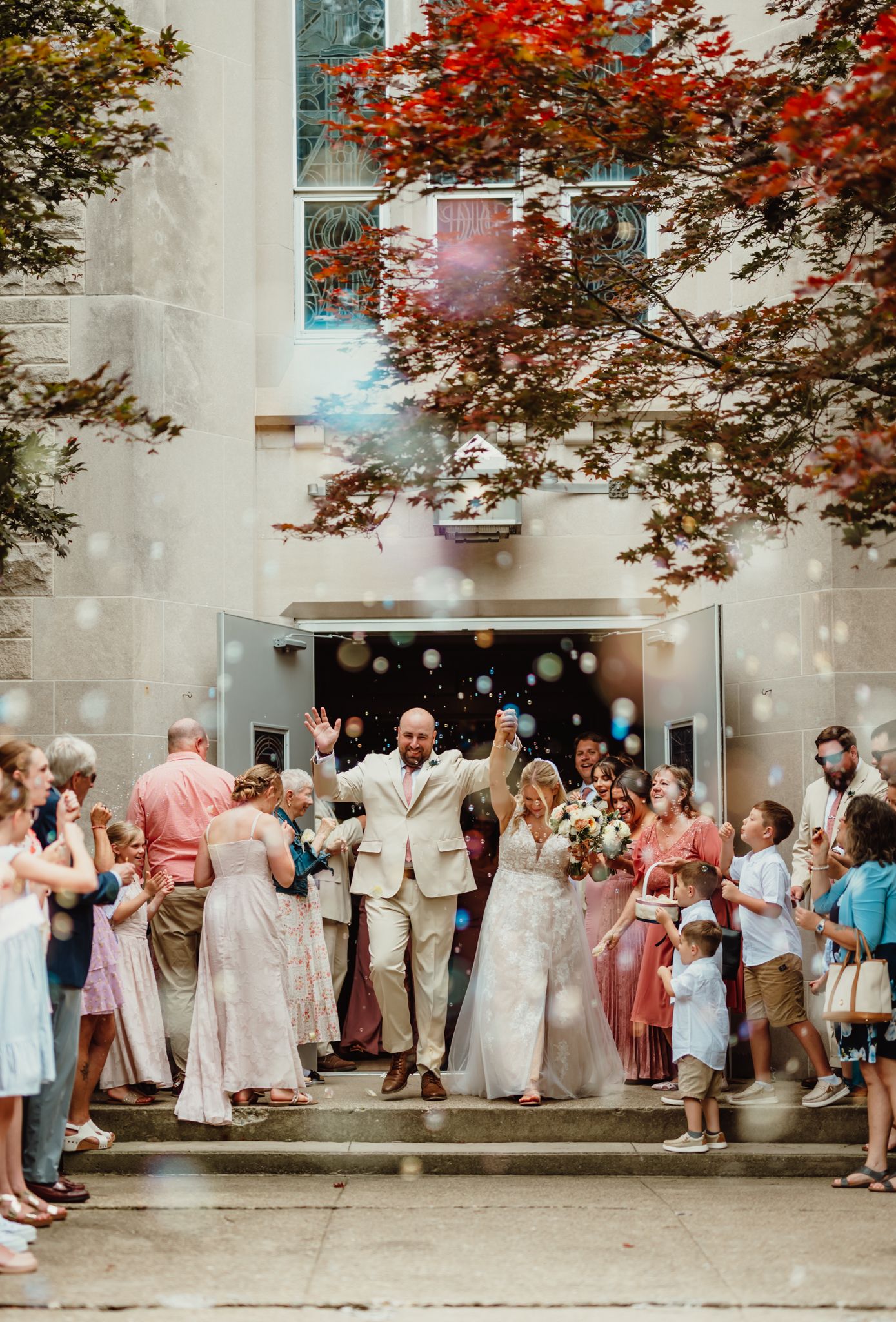 Newlywed couple exits church doors amid colorful confetti, surrounded by cheering guests on sunny wedding day.