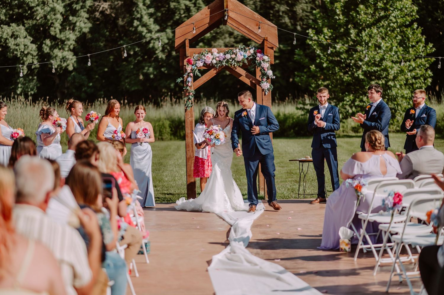 Officiant reads vows as bride and groom stand under floral wooden arch at outdoor ceremony.